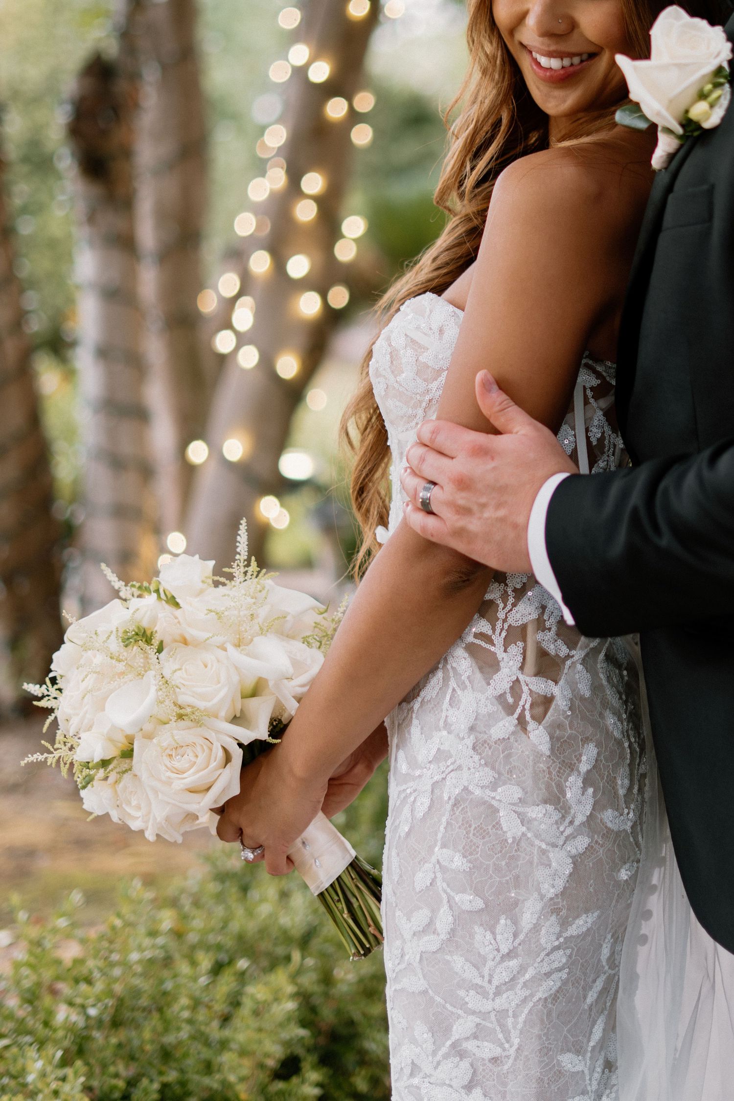 Groom embracing bride in lace wedding dress while she holds a white rose bouquet with bokeh fairy lights behind.