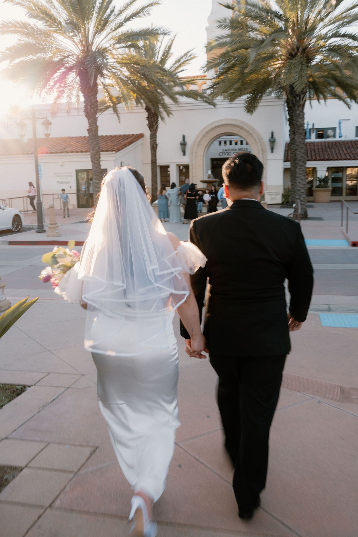Bride and groom walking hand in hand toward a venue surrounded by palm trees at golden hour.