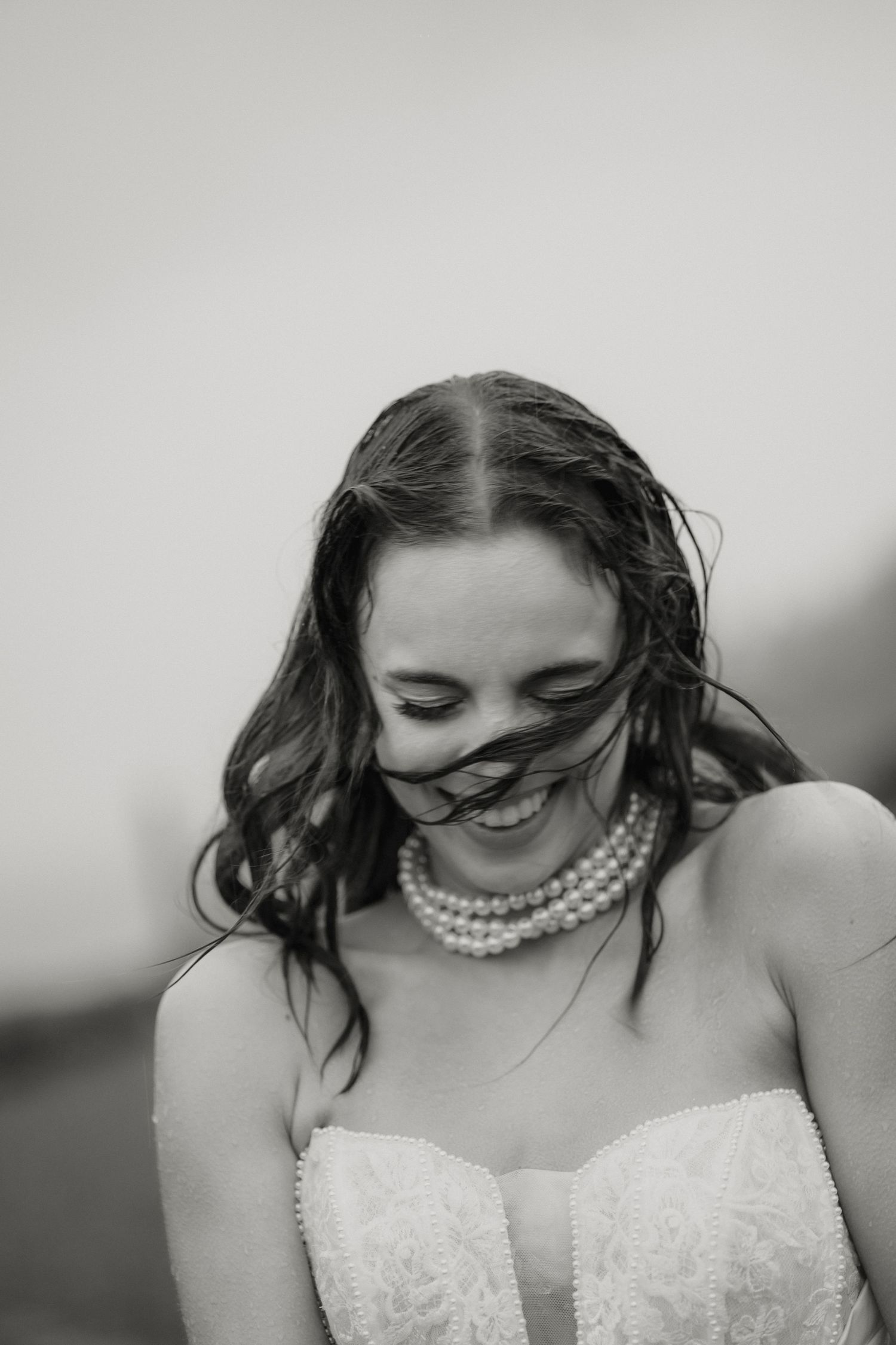 Black and white portrait of radiant bride smiling with wind-blown hair, wearing pearl necklace and strapless lace gown.