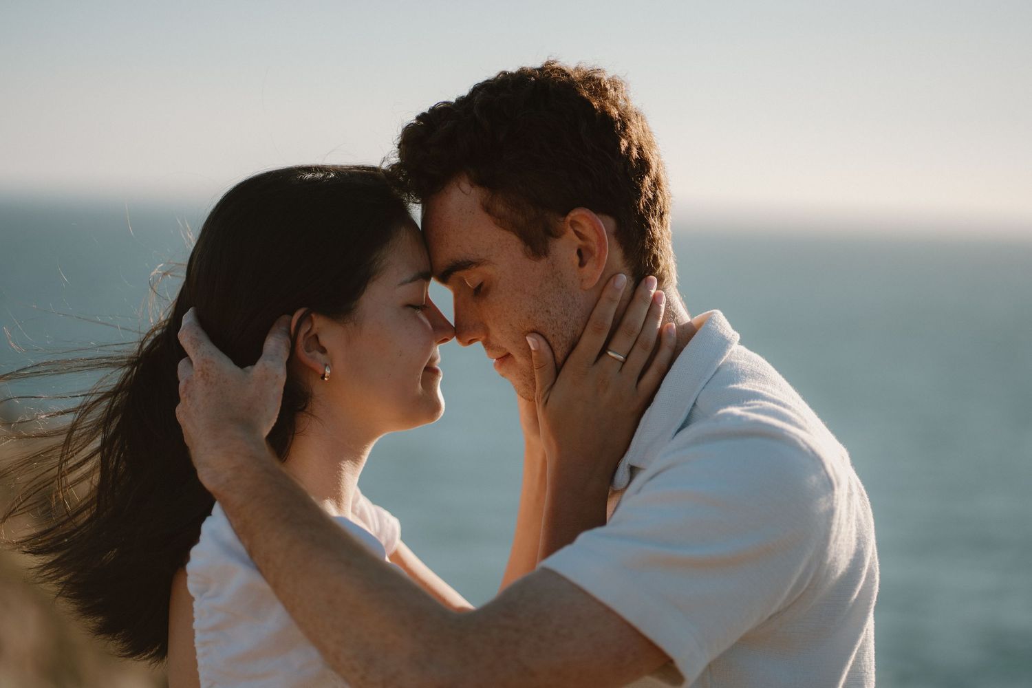 Romantic couple sharing an intimate moment with foreheads touching against ocean backdrop.
