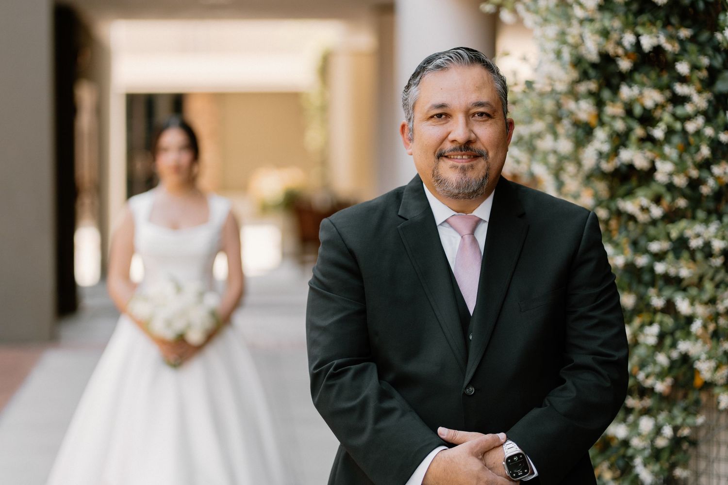 A man in a dark suit stands smiling with a bride in white holding a bouquet softly blurred behind him.