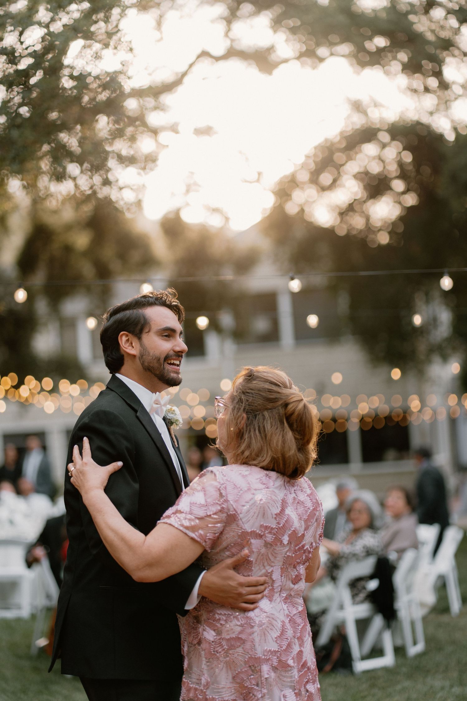 Groom shares a tender mother-son dance outdoors at golden hour wedding reception with string lights.