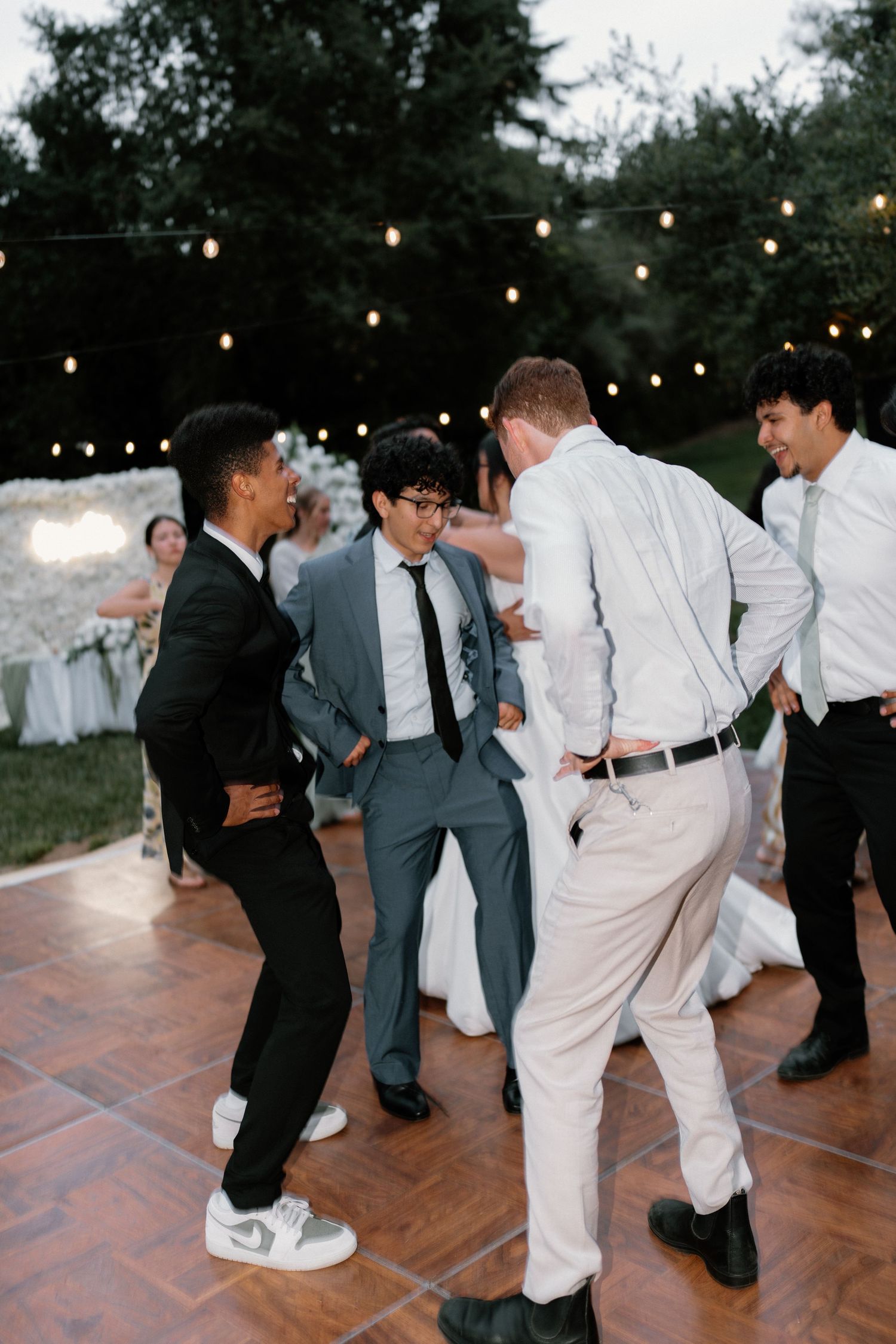 Wedding guests in suits dance and celebrate together on outdoor dance floor under string lights at night.