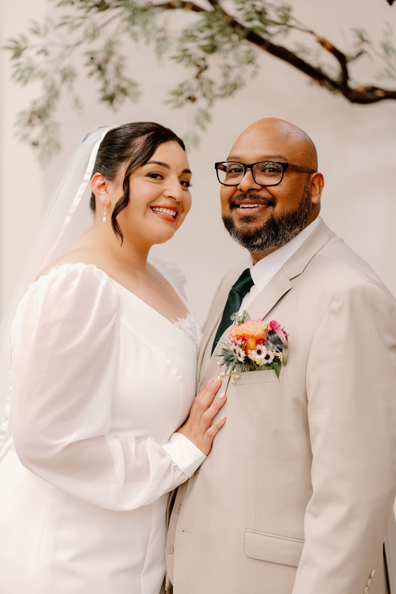 Smiling bride in white long-sleeve gown and groom in tan suit with floral boutonniere pose together at wedding.