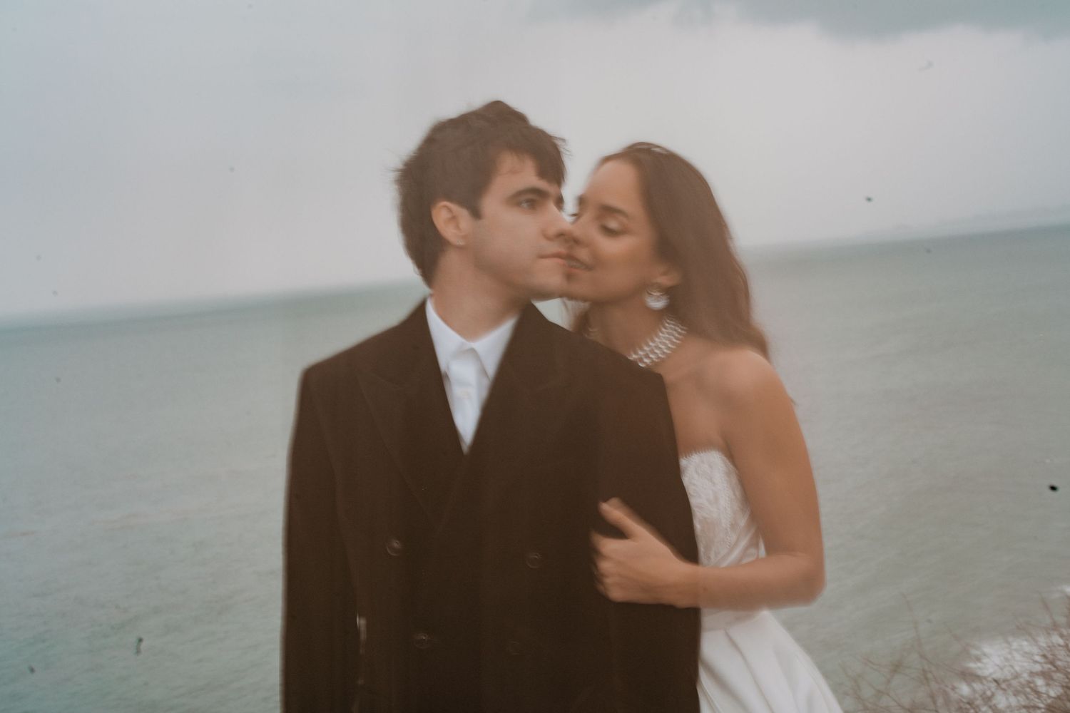 Bride kisses grooms cheek on overcast beach, she wears white strapless dress and he wears a dark suit.