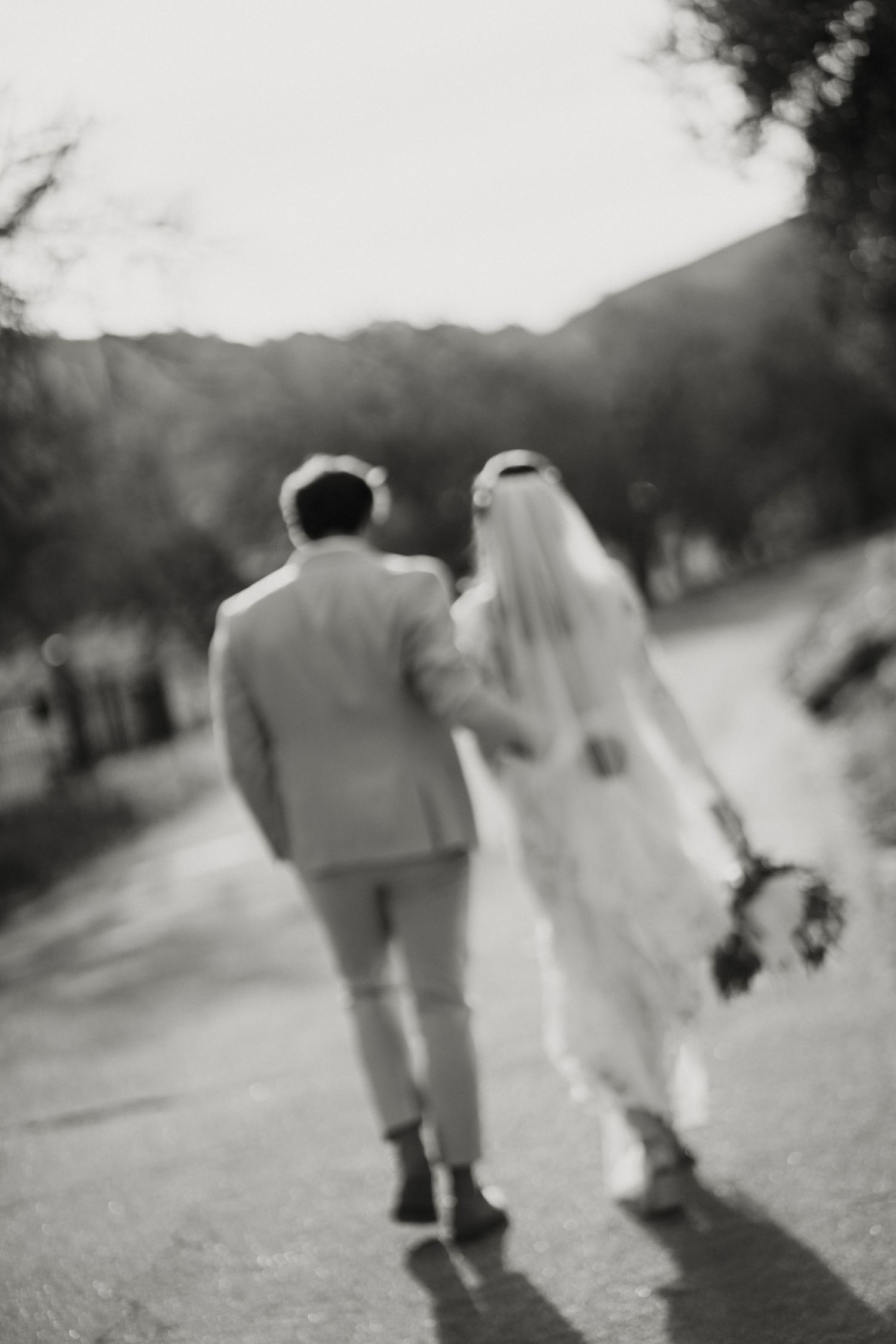 Artistic blurred black and white photo of bride and groom walking away down a tree-lined road.