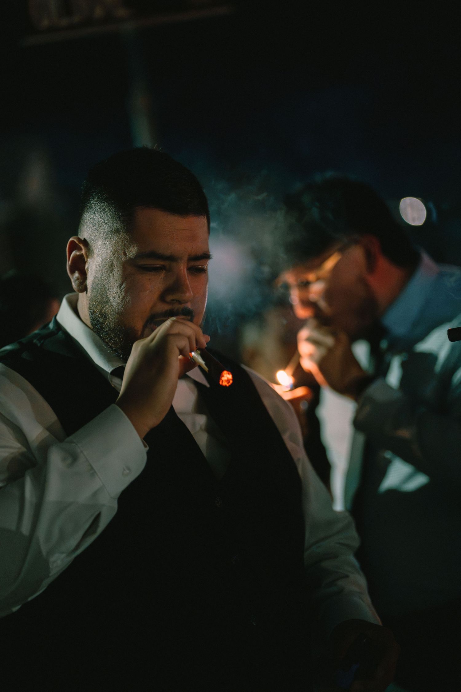 Groomsmen lighting cigars together in dim light during a wedding celebration.