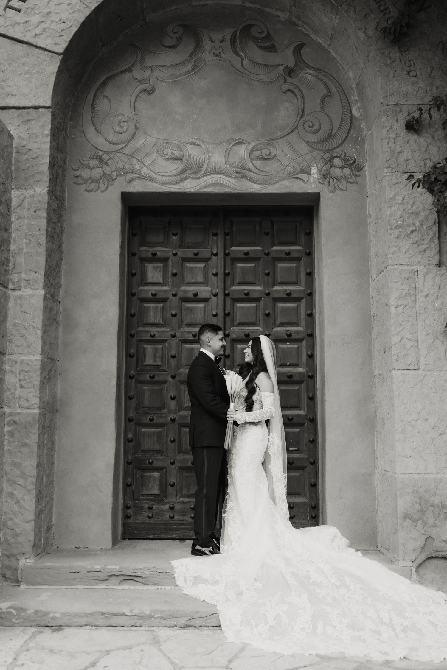 Black and white photo of bride and groom embracing before grand wooden doors of historic venue.