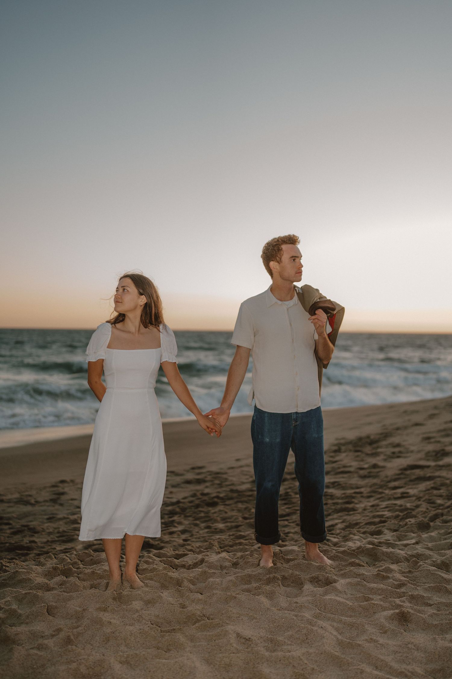 Couple holding hands on a beach at sunset, woman wearing white dress, man in white shirt and jeans, ocean in background.