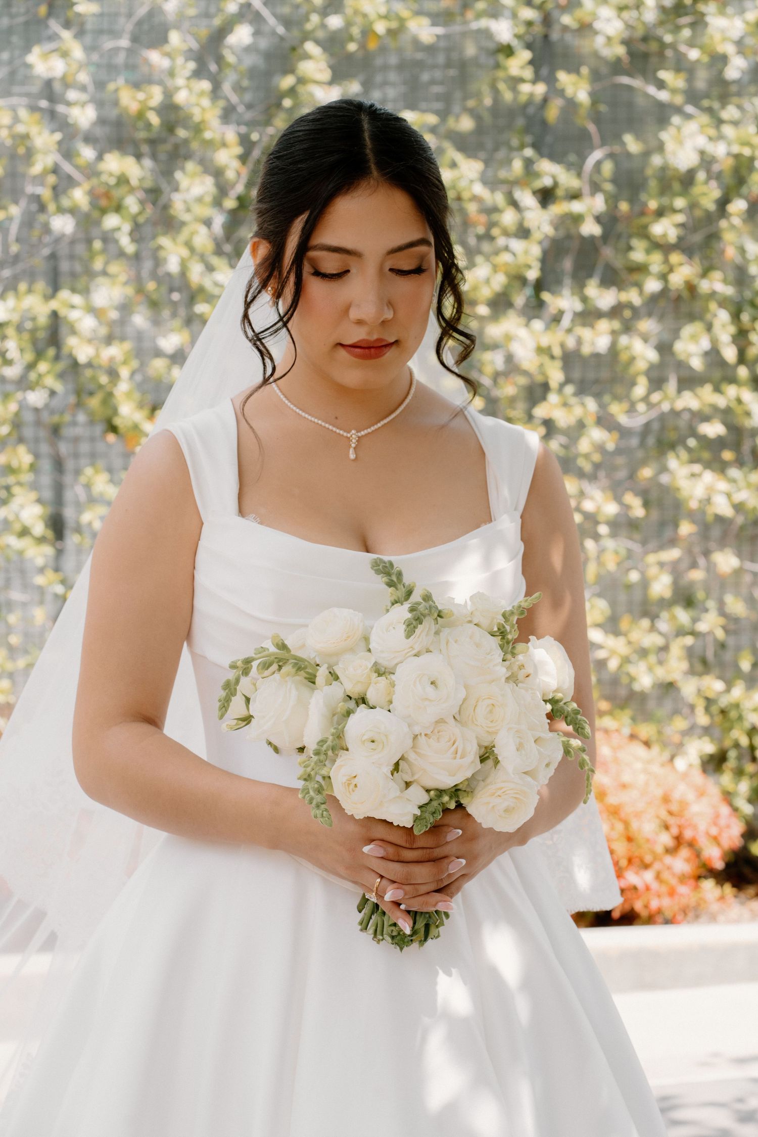 Beautiful bride in white square-neck gown holds white peony bouquet in front of flowering white trees.