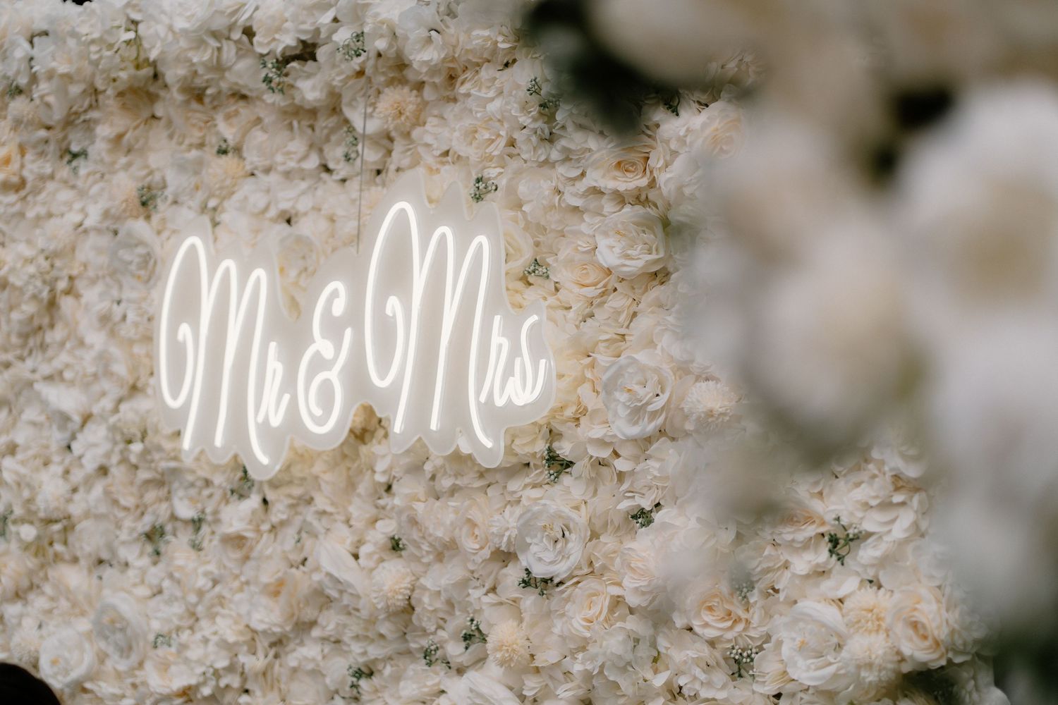 Elegant Mr & Mrs neon sign glows against a lush white floral wall backdrop at a wedding reception.