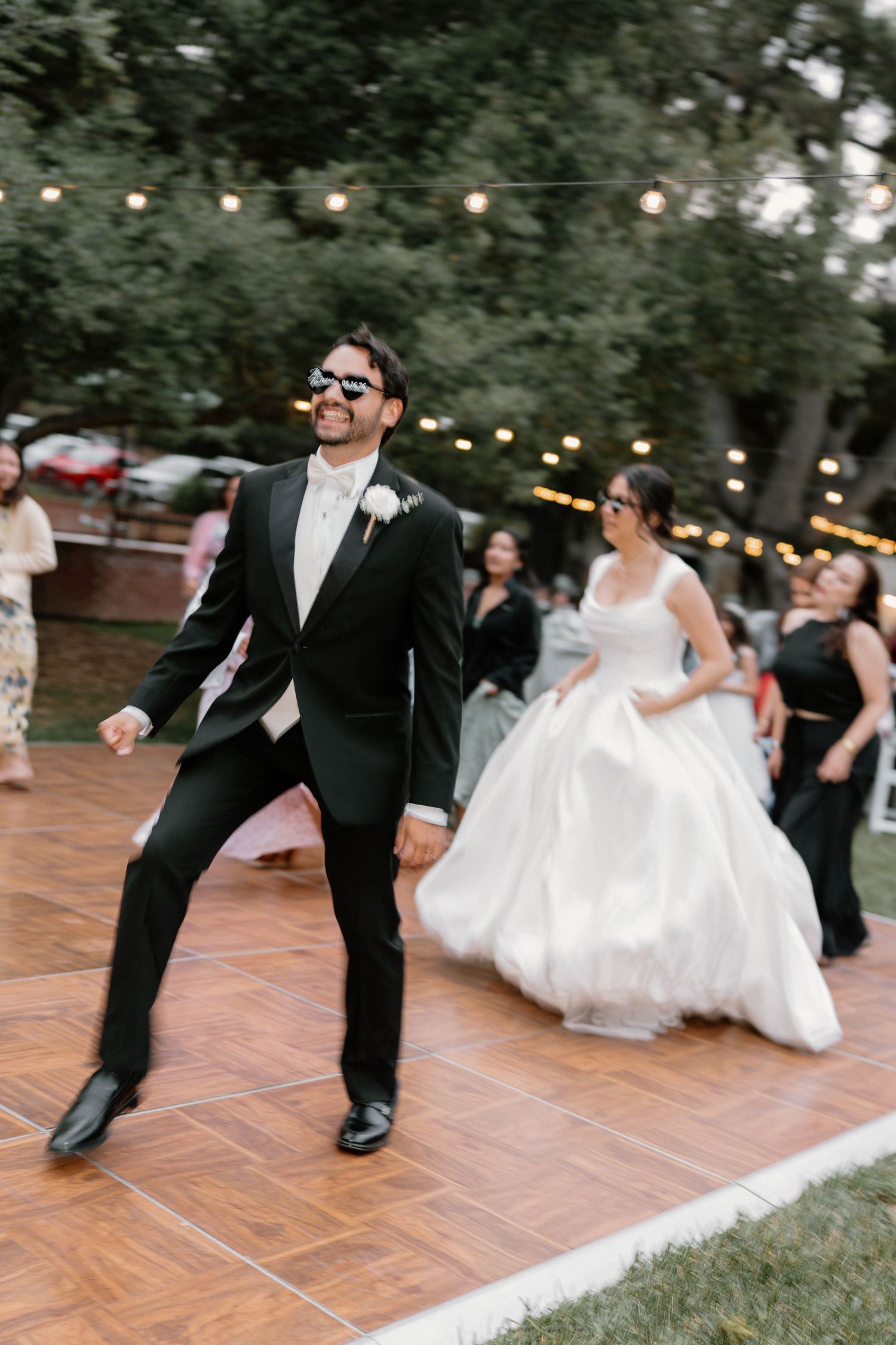 Groom dances energetically in heart sunglasses on outdoor dance floor at lively wedding reception.