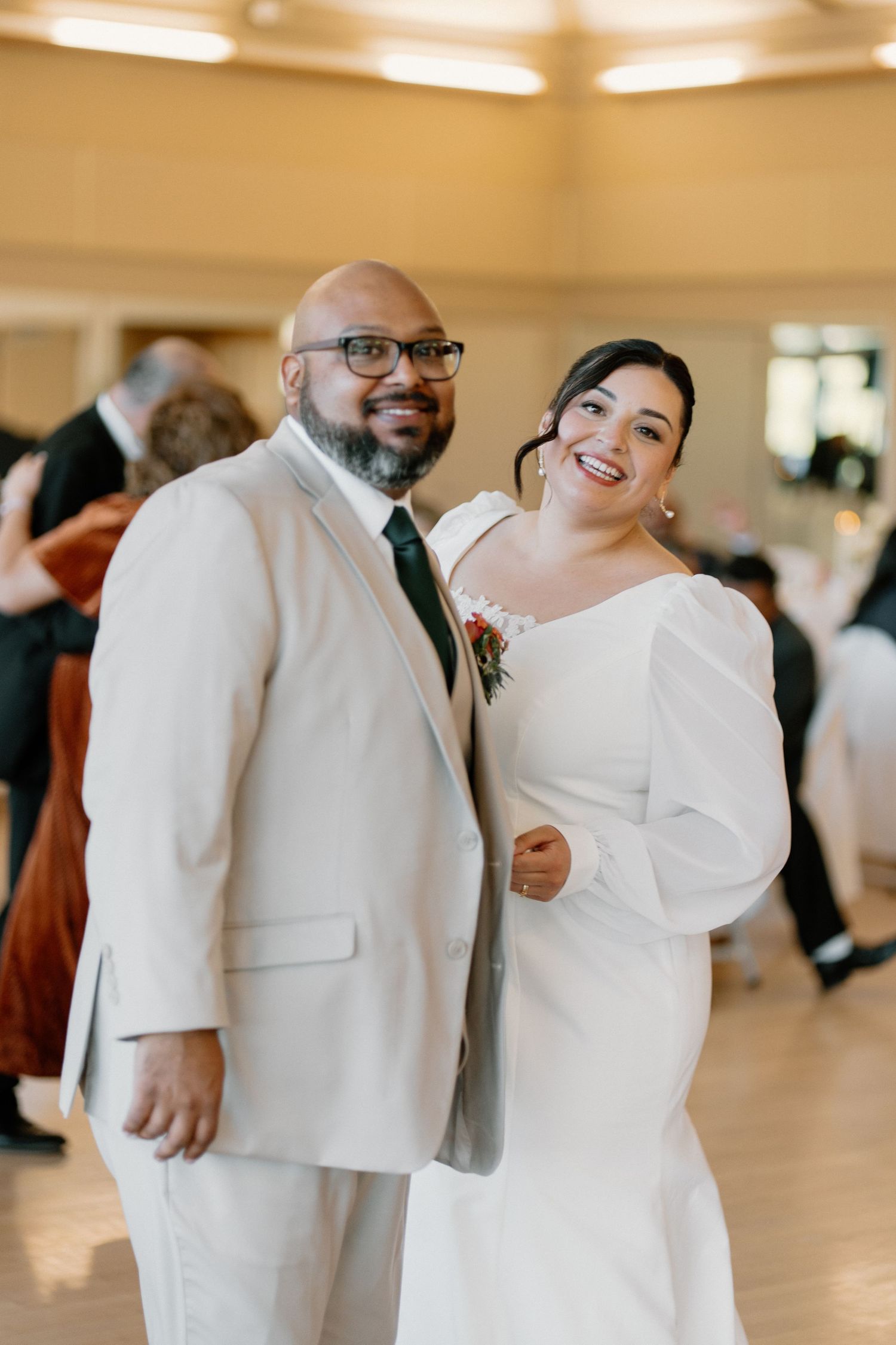 Smiling bride and groom in white dress and tan suit posing together on wedding dance floor.
