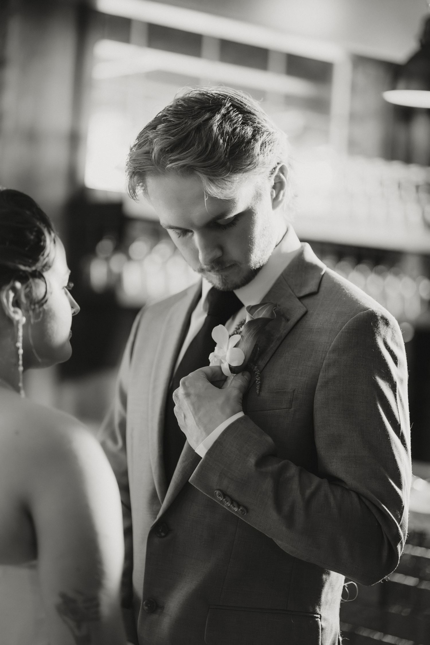 Groom in gray suit adjusting boutonniere while bride looks on in black and white wedding photo.