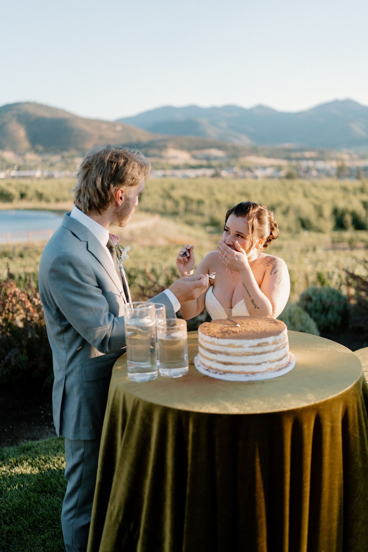 Bride and groom cutting naked wedding cake outdoors with scenic mountain and lake backdrop.