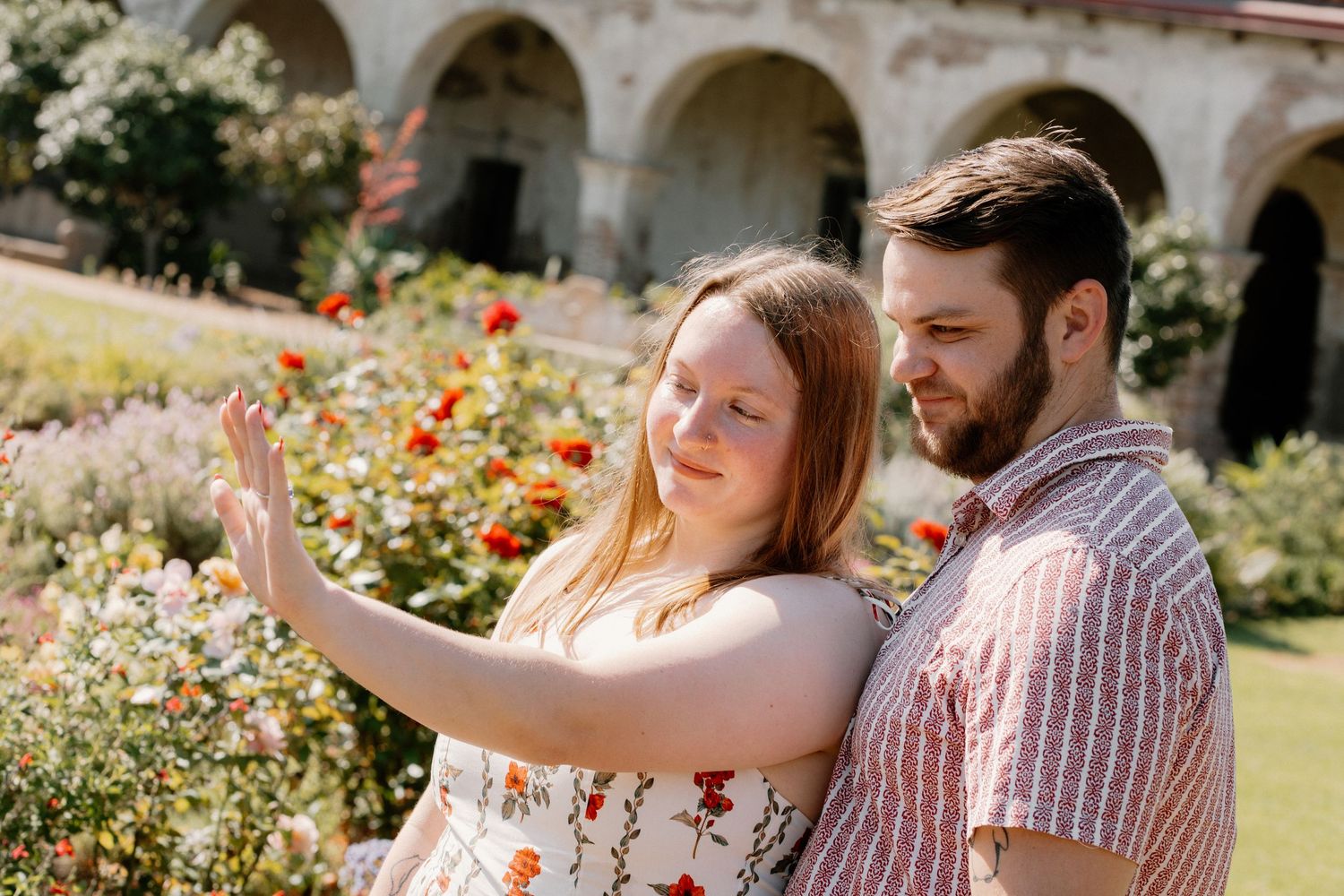Couple enjoying a sunny garden with blooming roses, woman reaching toward flowers while man stands close beside her.
