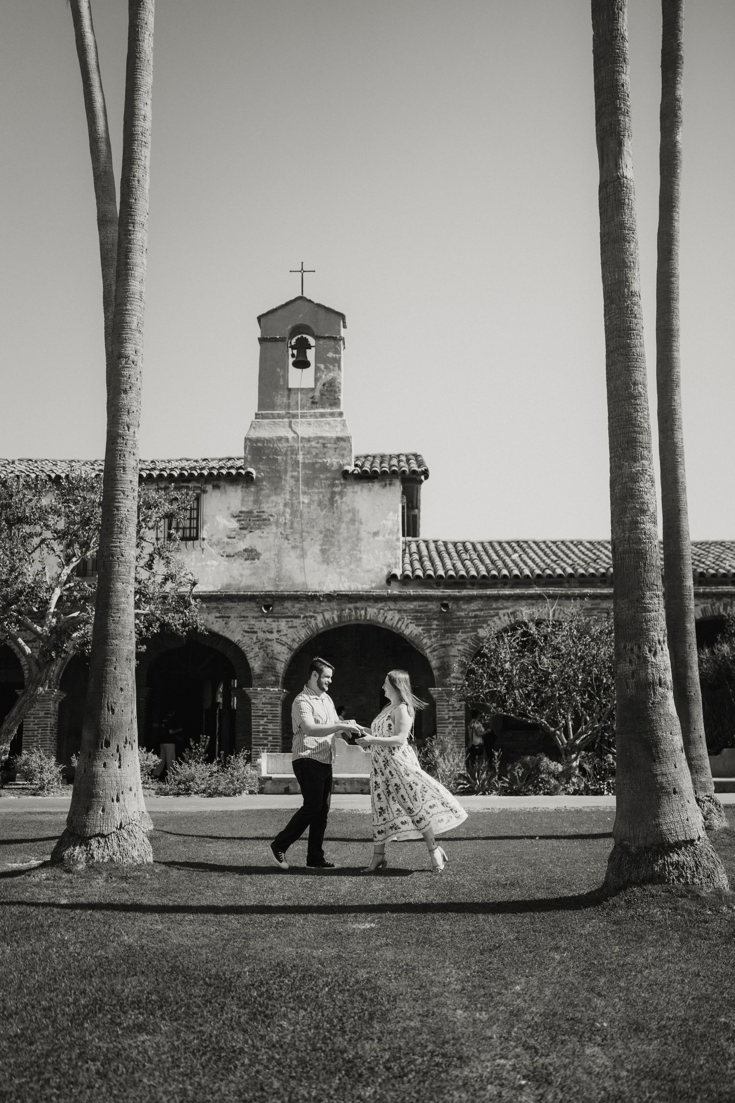 Black and white photo of couple dancing on lawn before historic mission bell tower framed by tall palm trees.