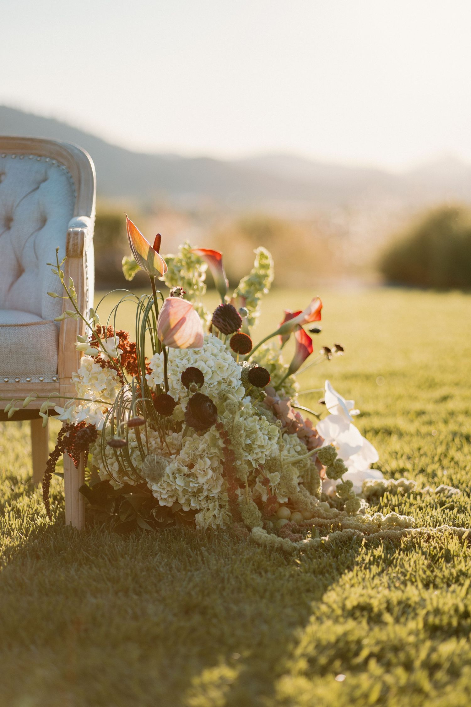 Lush bridal bouquet with tropical flowers resting against a vintage tufted chair on a green lawn.