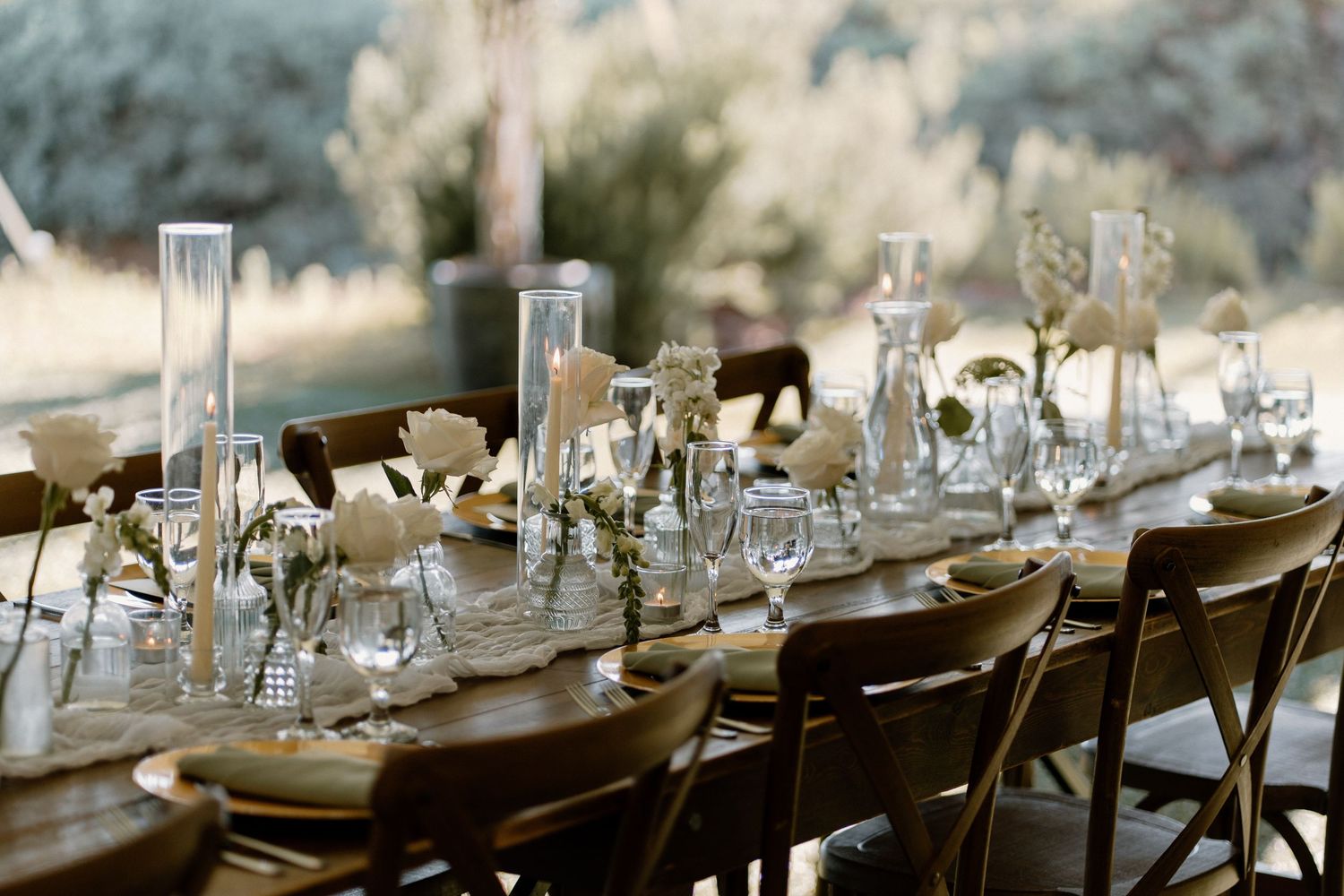 Elegant outdoor wedding reception table with white roses, candles, and glassware centerpieces.