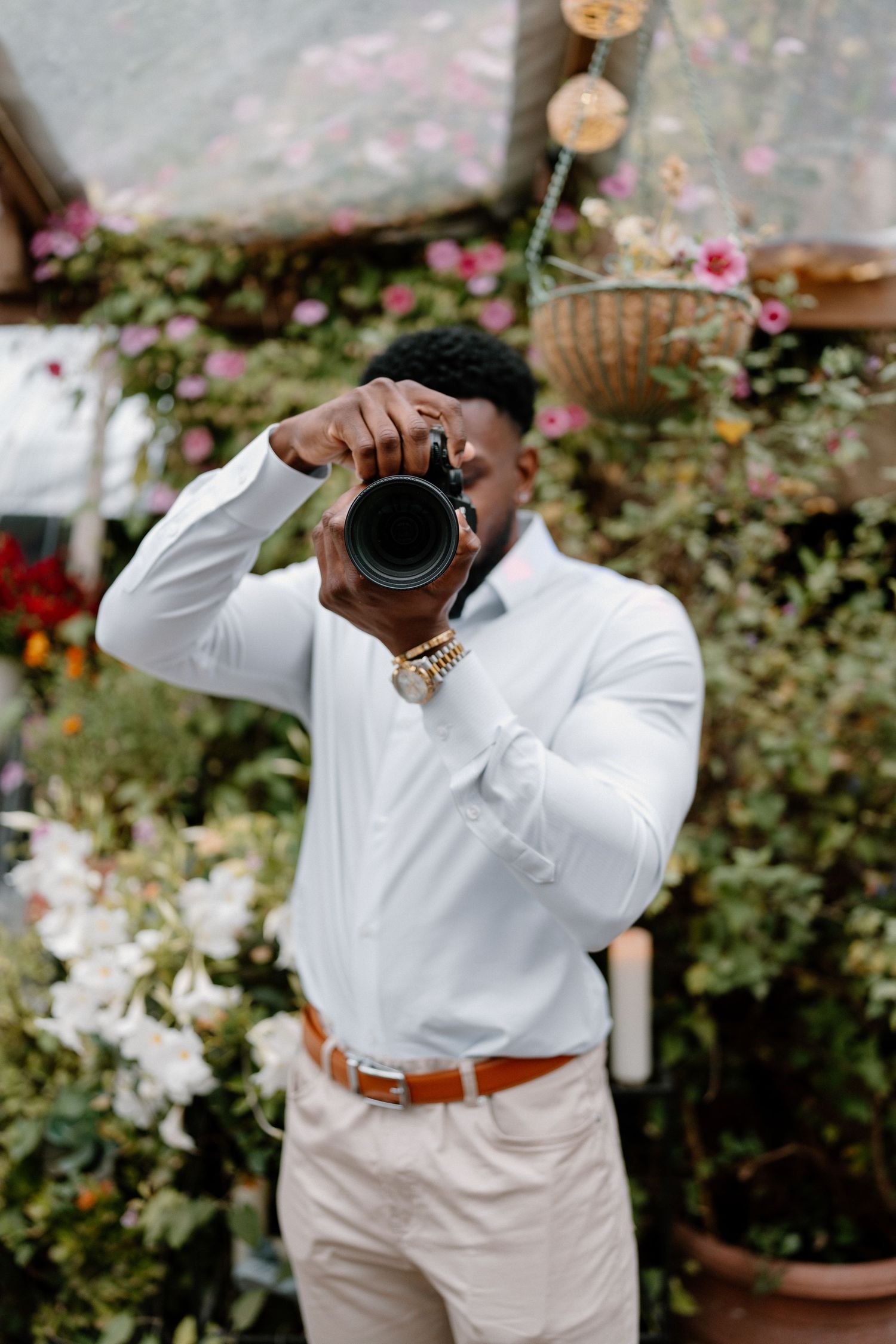 Stylish wedding photographer in white shirt aims camera toward viewer amid a lush floral garden backdrop.