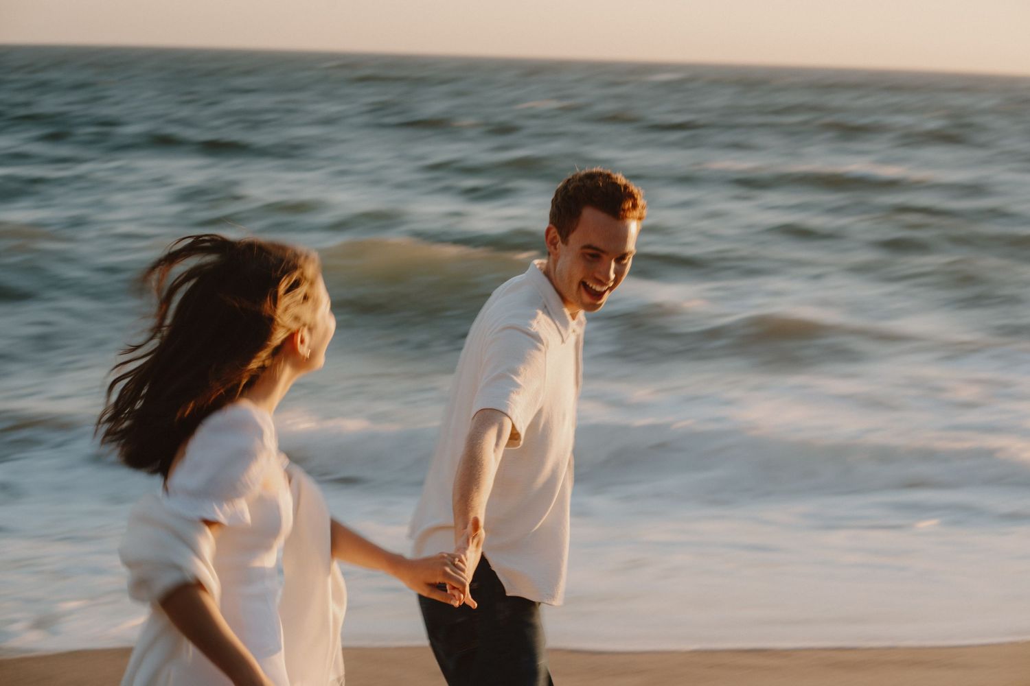 Happy couple holding hands and walking along a sandy beach at golden hour by the ocean.