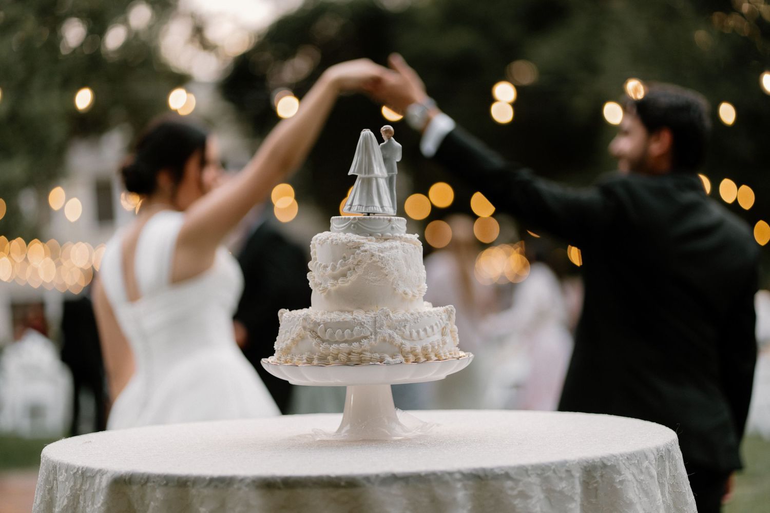 Elegant wedding cake with bride and groom topper in foreground as couple dances at outdoor reception.