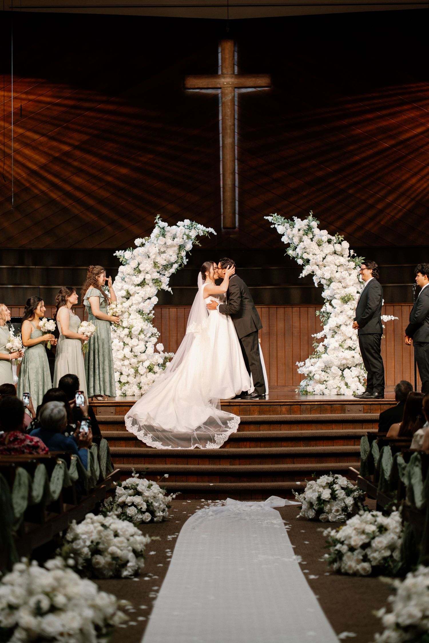 Newlyweds share first kiss under floral arch in church with bridesmaids and groomsmen present.