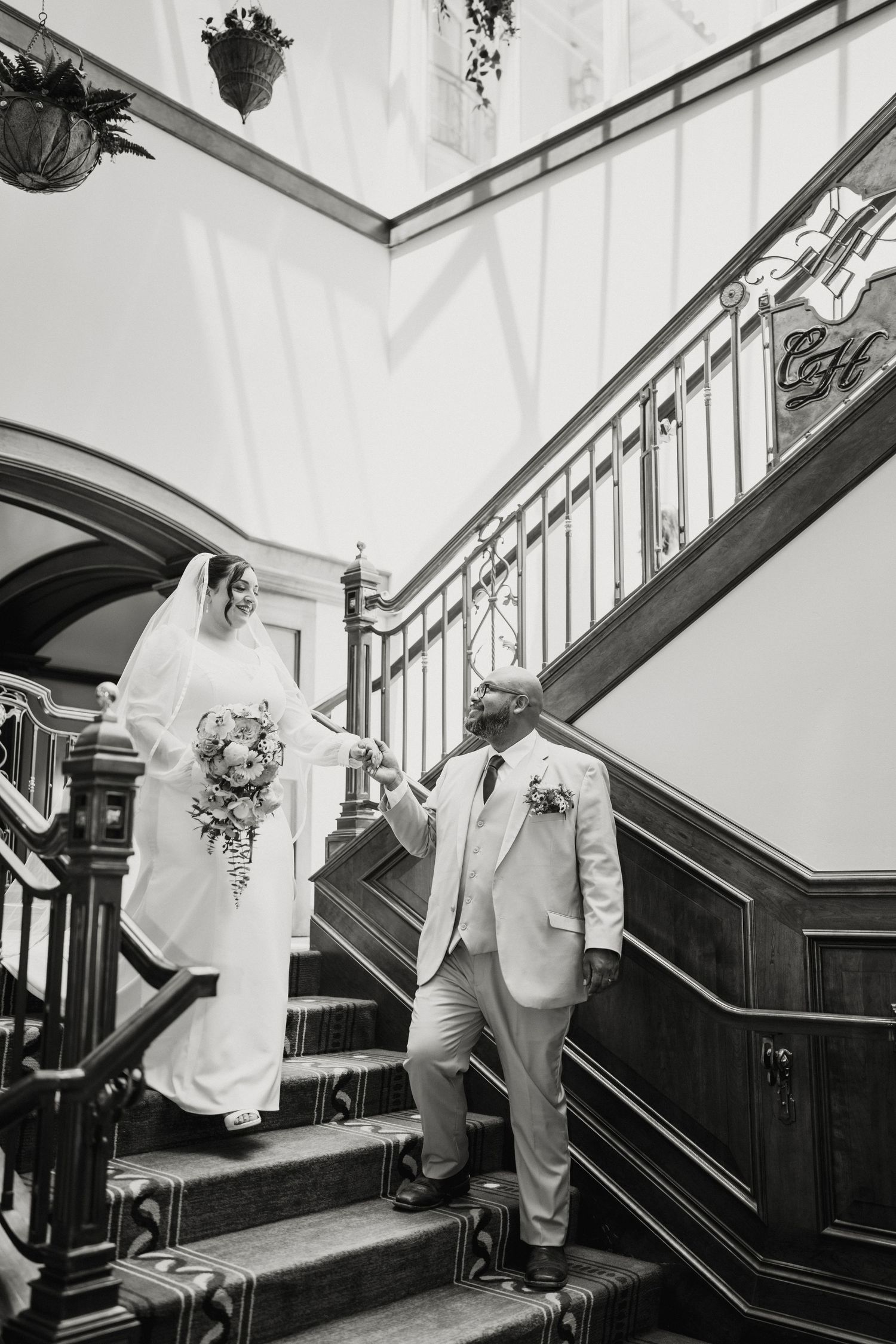 Black and white photo of bride and groom descending elegant wooden staircase at wedding venue.