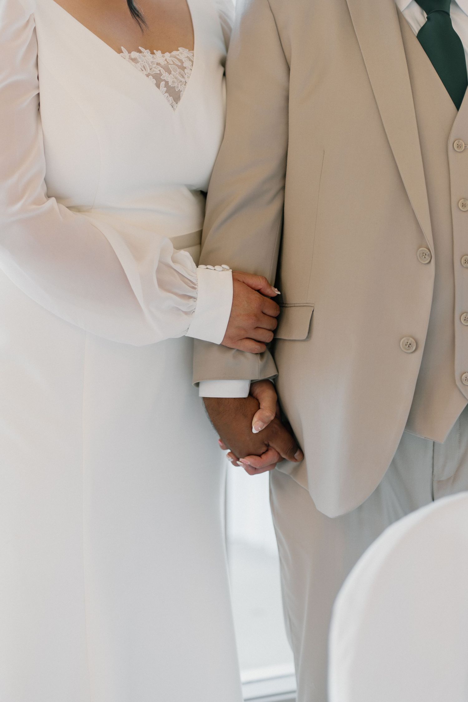 Bride and groom holding hands at wedding ceremony, bride in white lace dress, groom in beige suit.