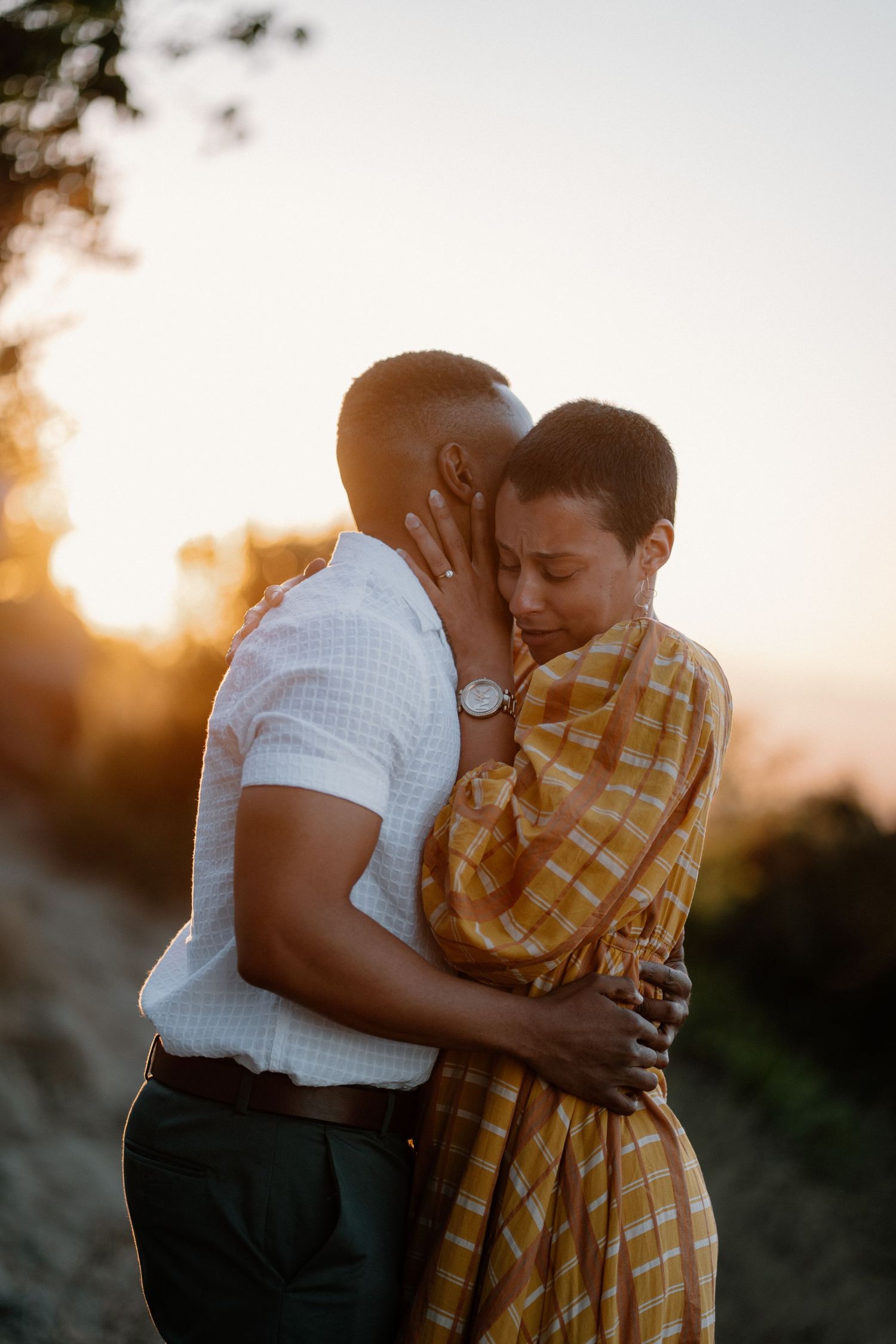 Couple sharing a tender embrace at golden hour sunset, woman in yellow plaid dress and man in white shirt.