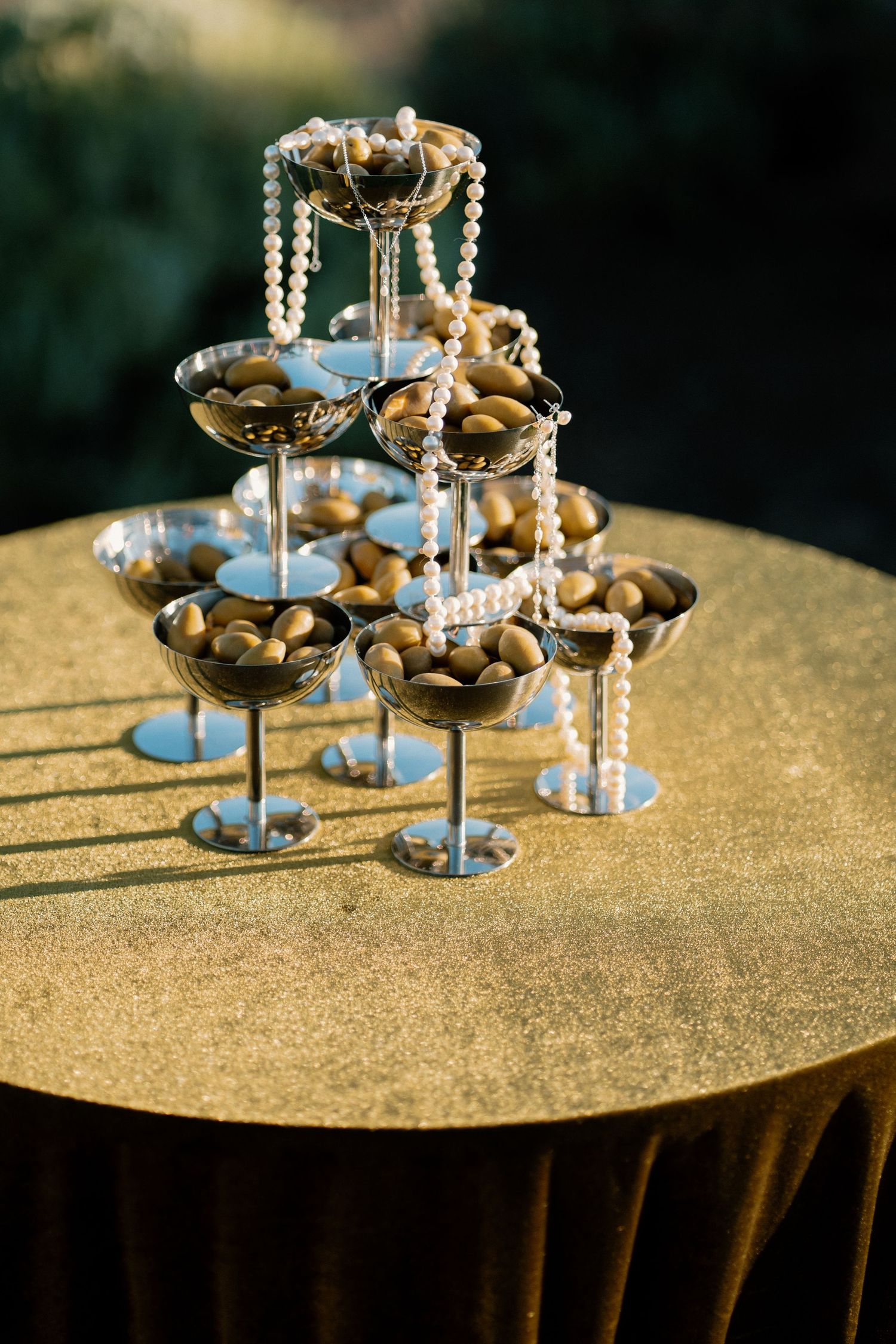 Champagne tower with pearl garlands and gold-filled coupe glasses on a gold tablecloth outdoors.