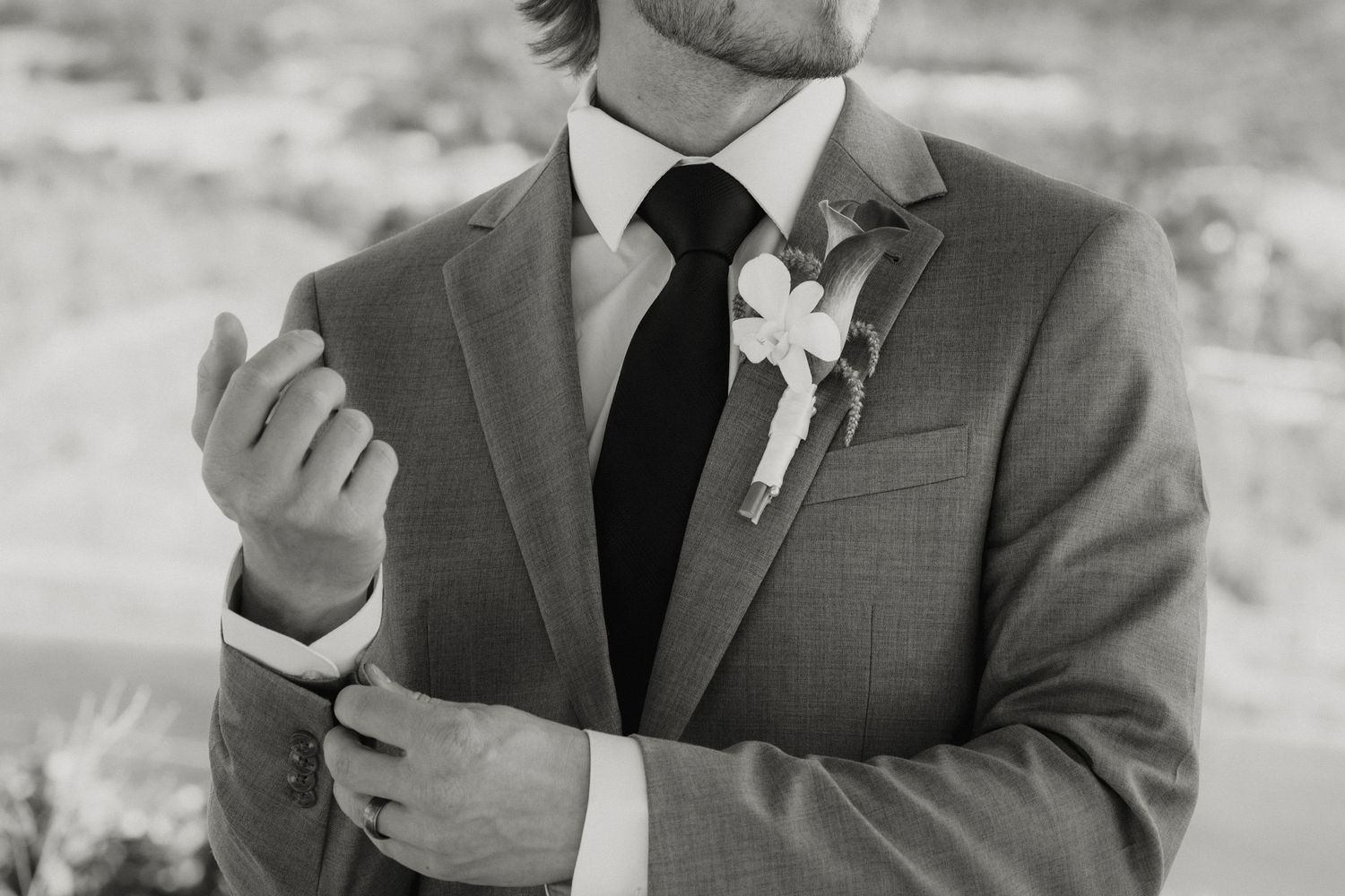 Groom in gray suit adjusting cufflinks with boutonniere on lapel, black and white wedding photo.