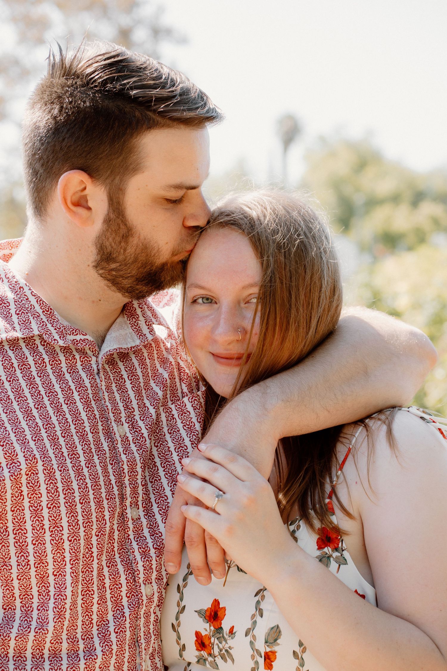 Bearded man kissing redhead woman on temple while embracing her from behind outdoors.