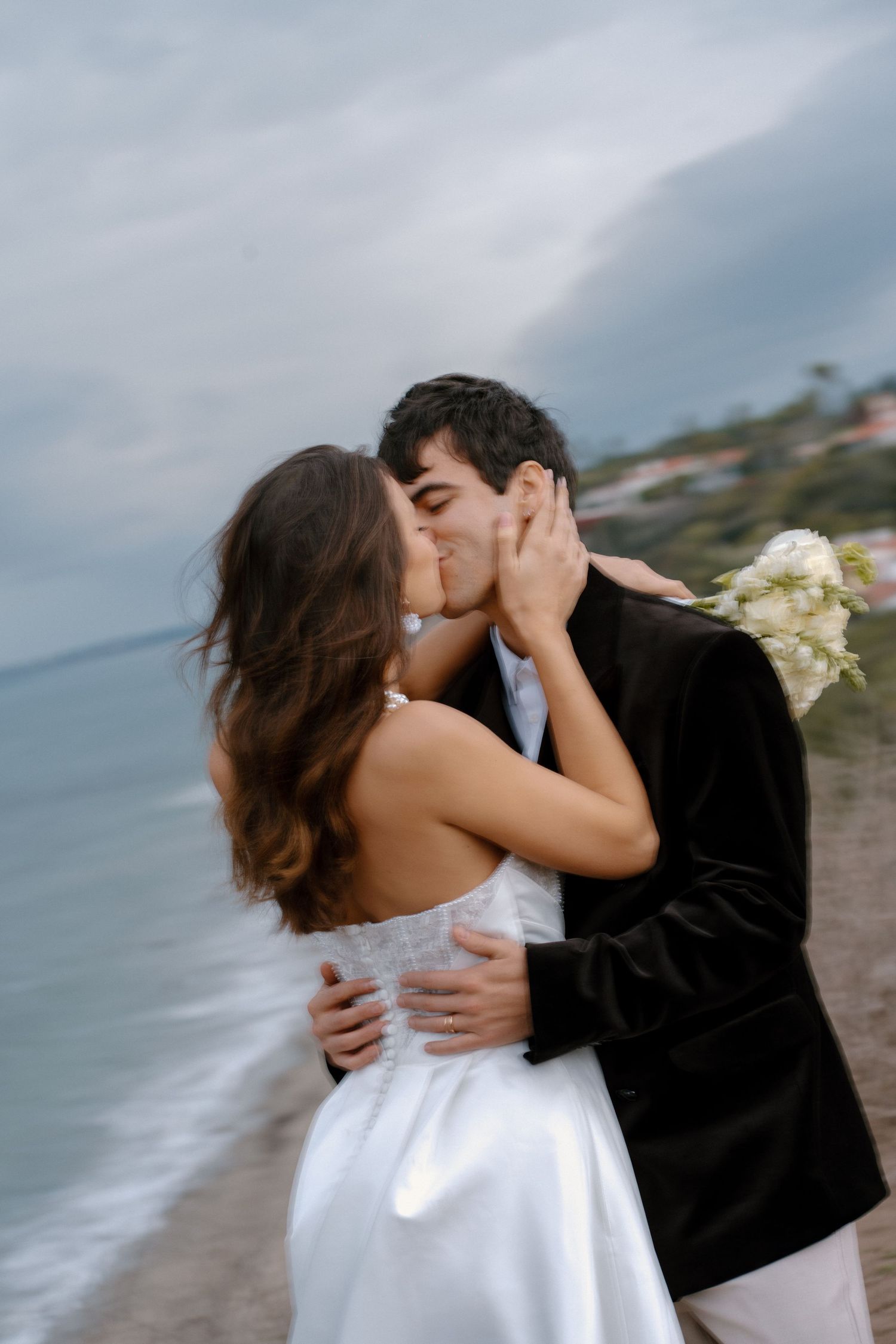 Bride and groom share romantic kiss on beach with bouquet of white flowers.