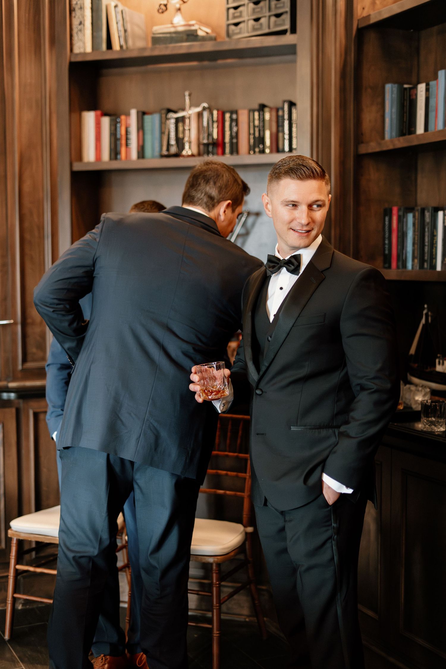 Dapper groom in black tuxedo smiles confidently in wood-paneled library bar during wedding celebration.