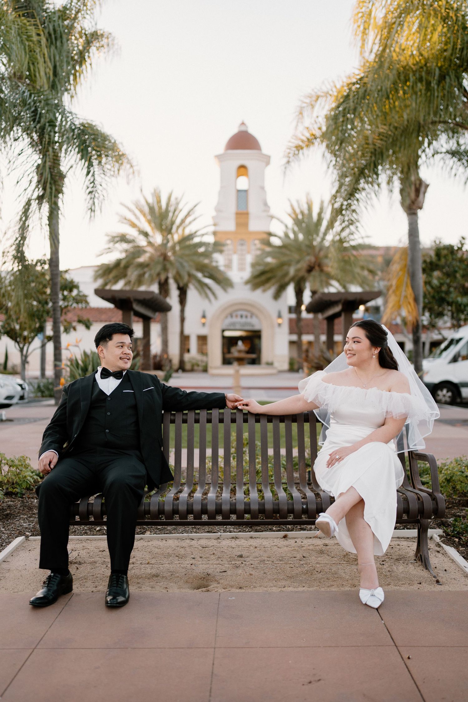 Bride and groom holding hands on a bench with palm trees and a church tower in the background.