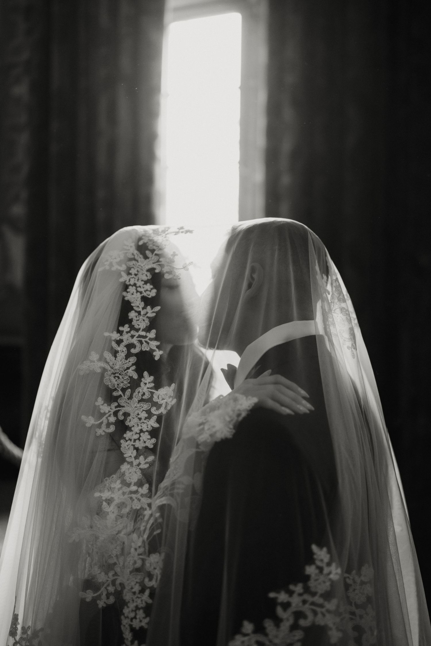 Black and white artistic photo of bride and groom kissing beneath lace veil by bright window.