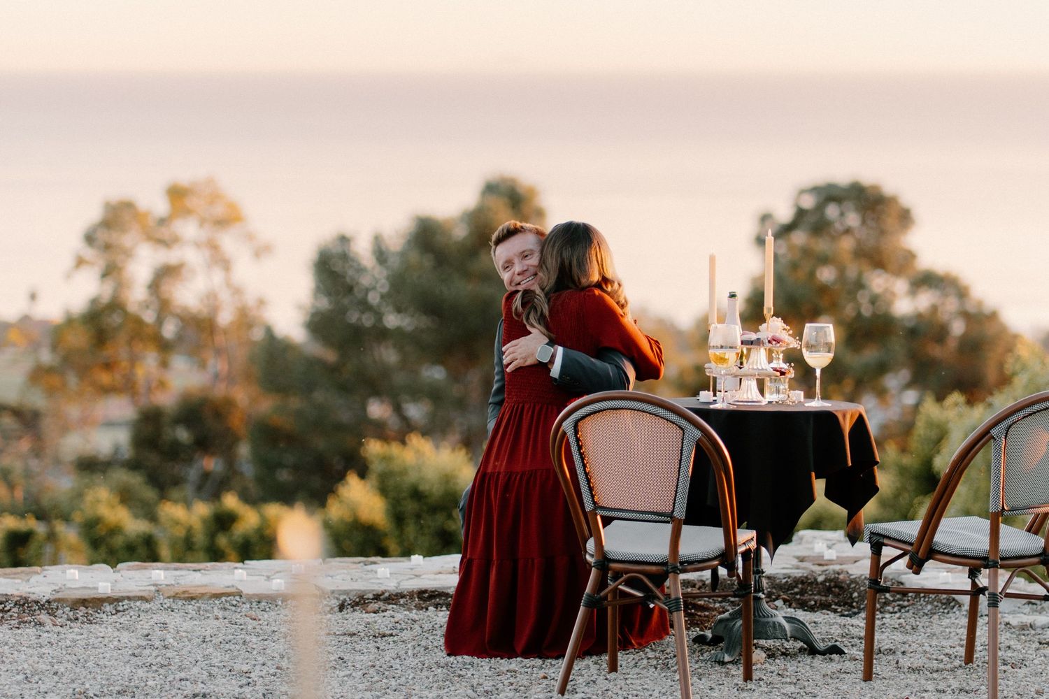 Couple embraces at a candlelit outdoor table overlooking the ocean at sunset.
