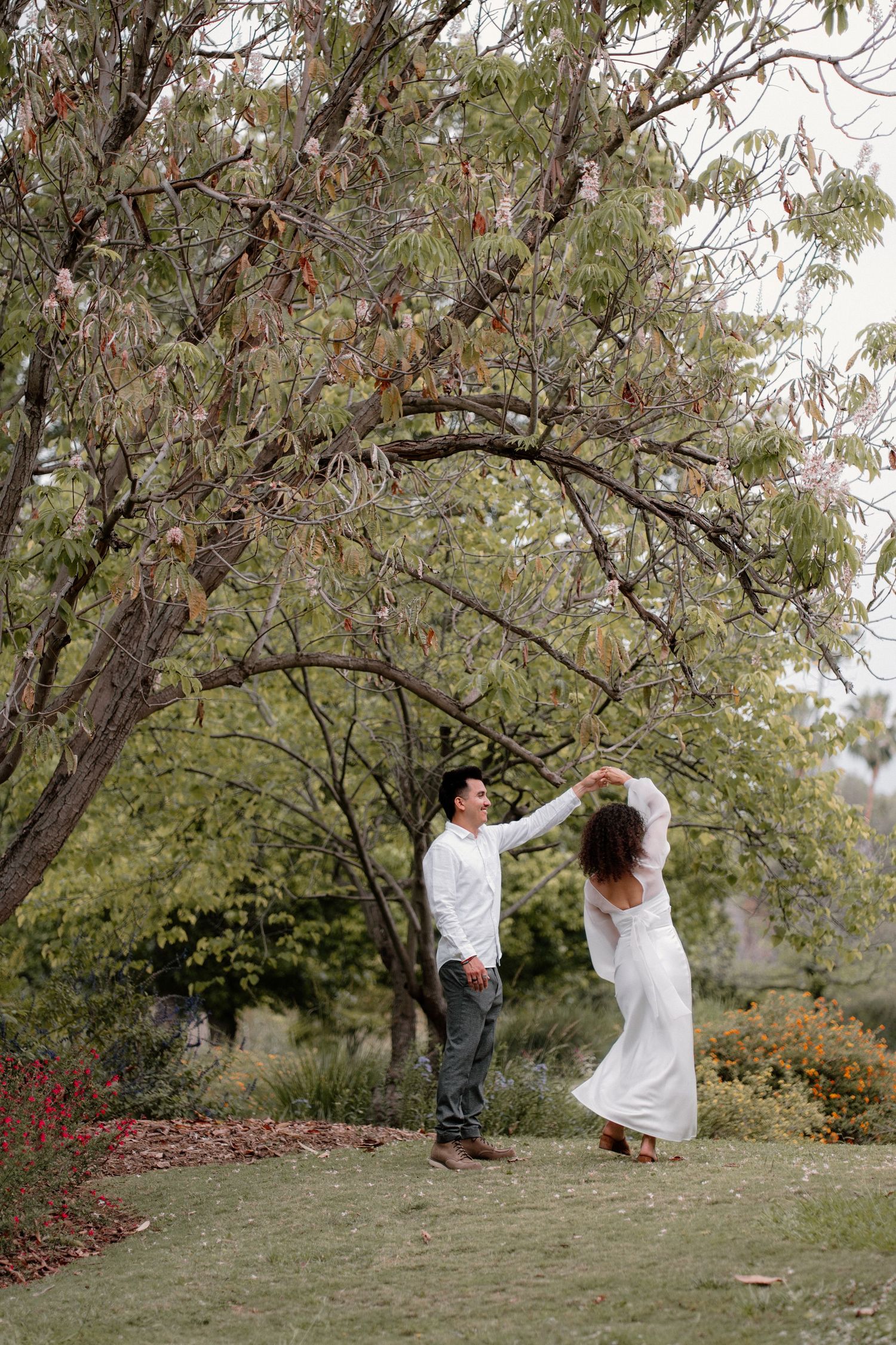 Couple dancing under a tree in a garden, bride in white dress being twirled by groom in white shirt.