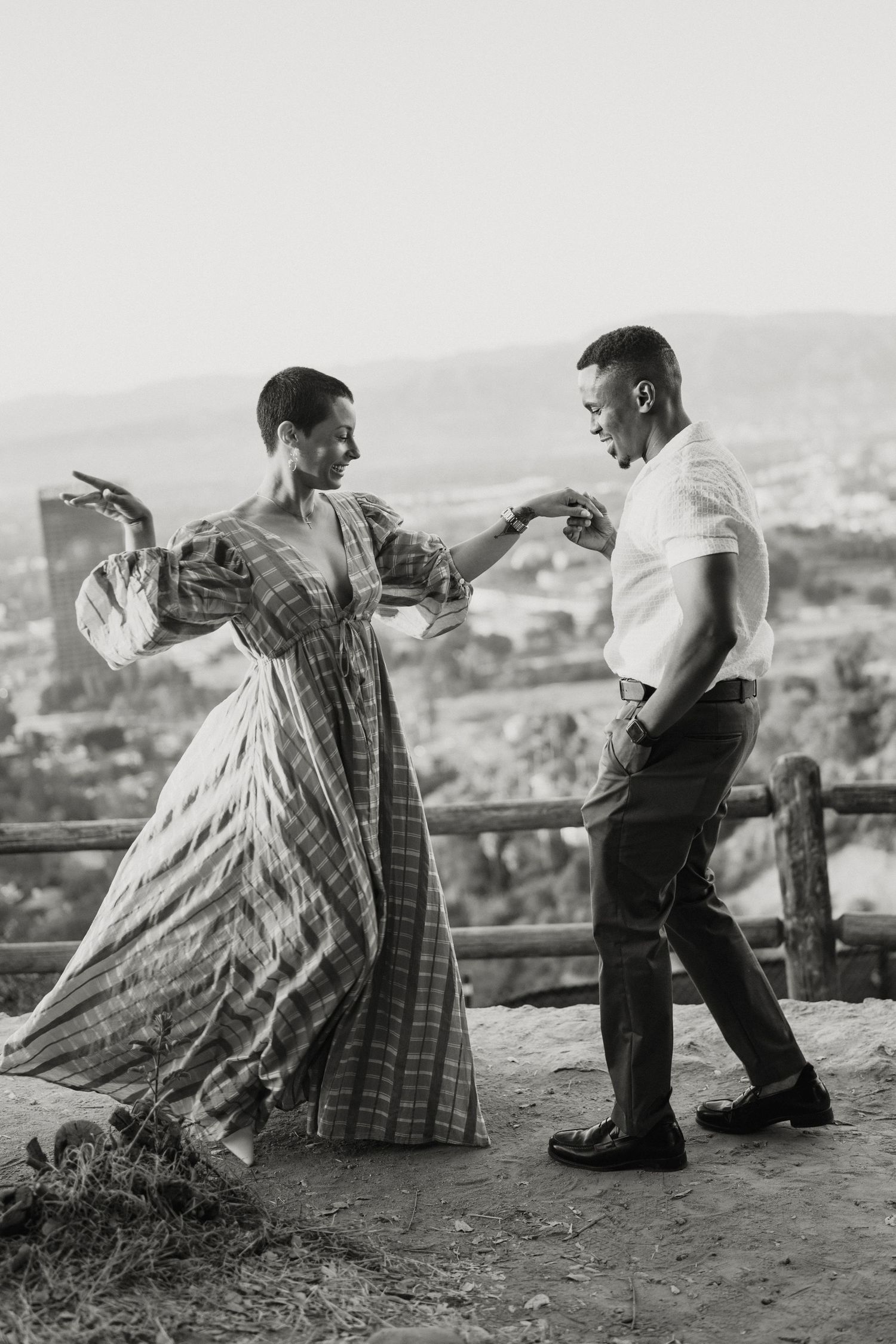 Black and white photo of a couple dancing joyfully at an outdoor overlook with a city view.