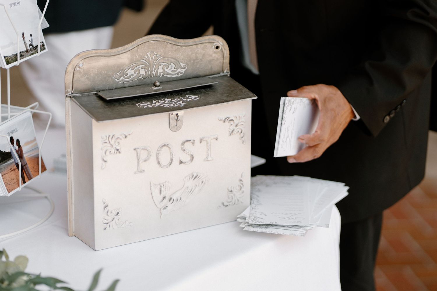 Groomsman placing card into decorative silver POST mailbox on white table at wedding reception.