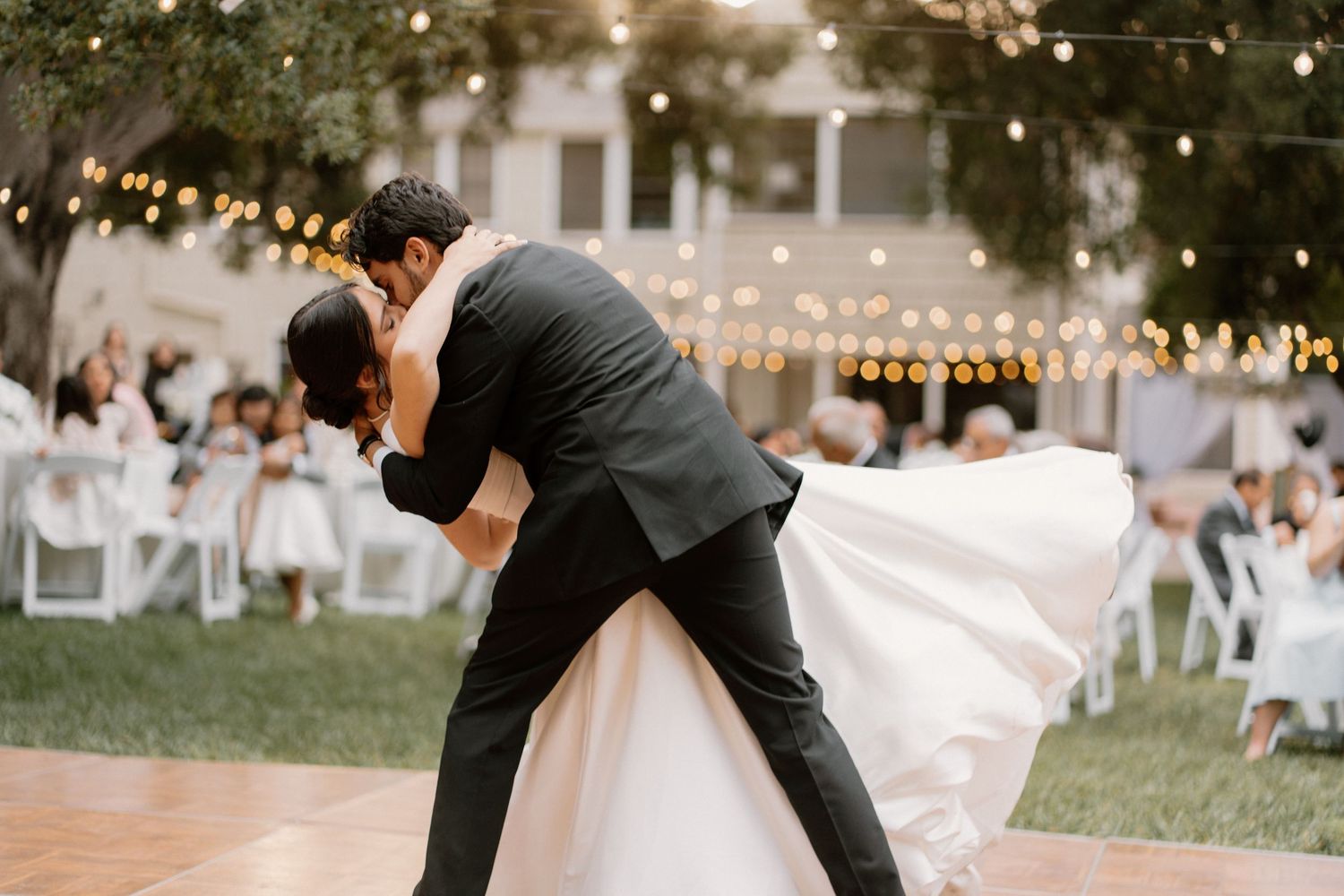 Groom dips bride in romantic first dance under string lights at outdoor wedding.