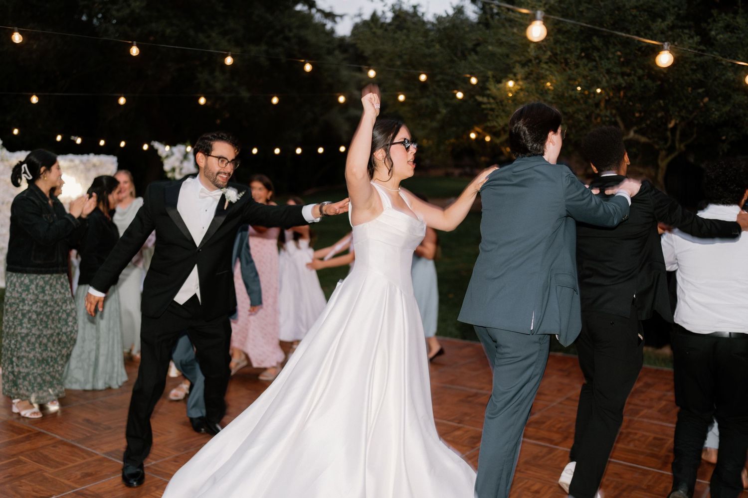 Bride in white ball gown dances joyfully with groom and guests at outdoor wedding reception under string lights.