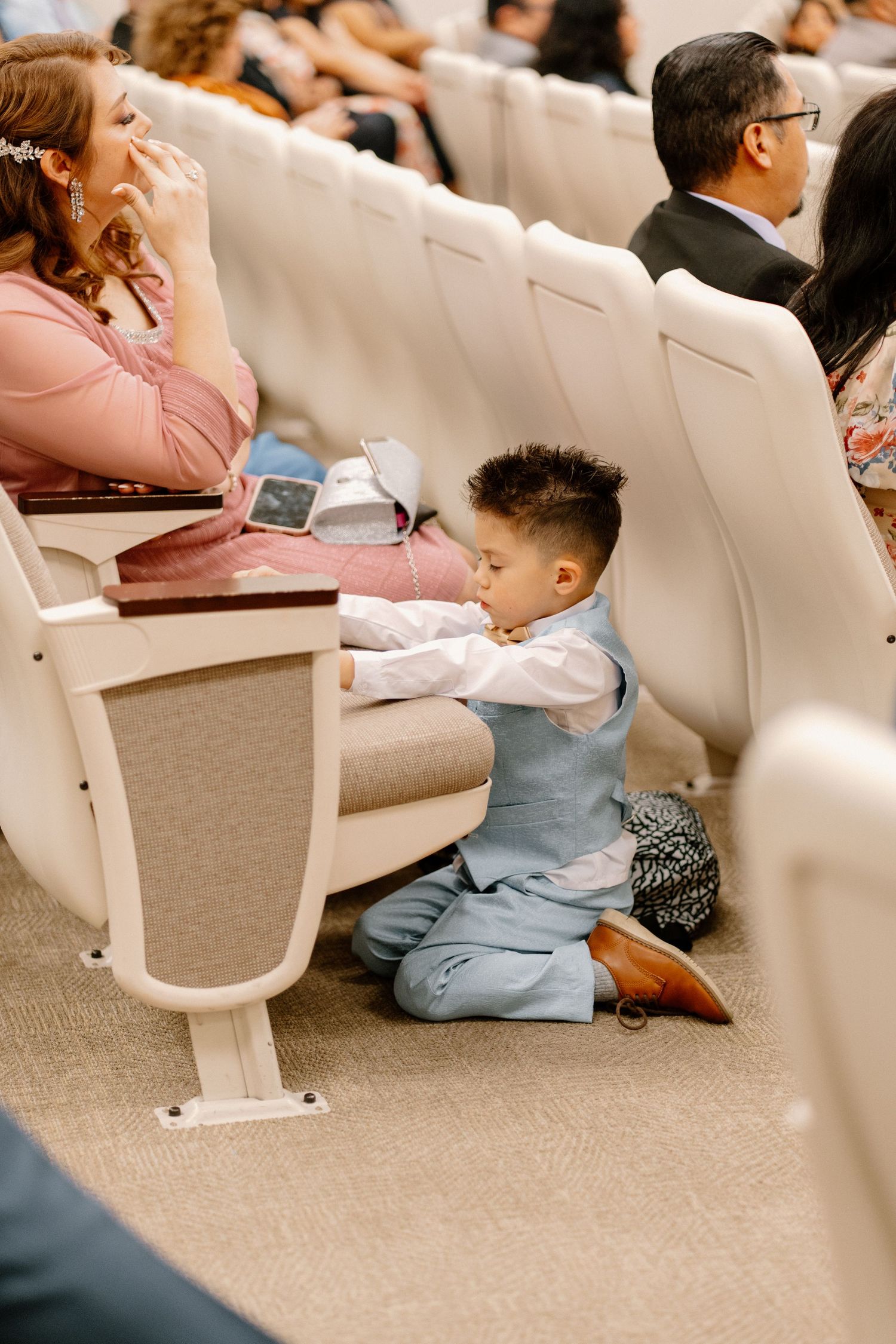 Young boy in blue vest sits on floor between chairs during a formal ceremony indoors.