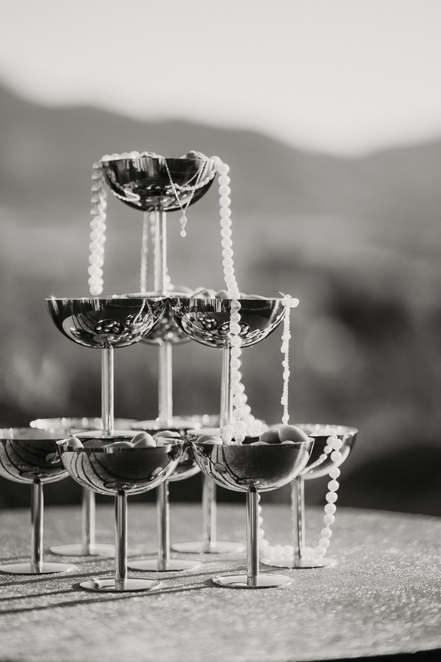 Black and white photo of elegant champagne tower with pearl necklace draped over coupe glasses.