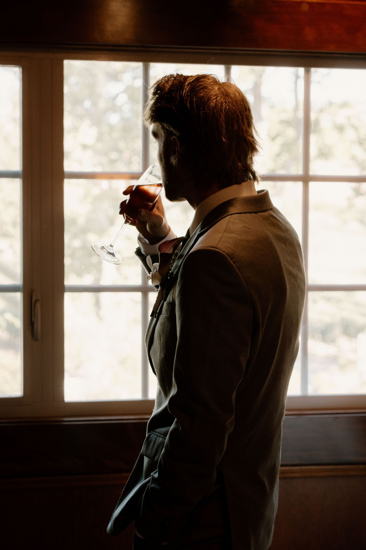 Silhouette of groom in gray suit sipping wine by sunlit window before wedding ceremony.