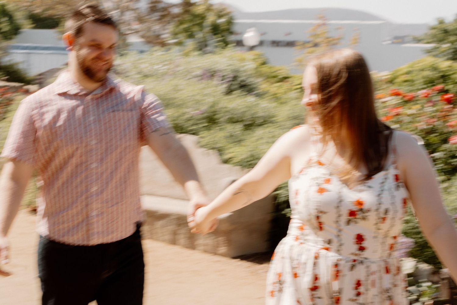 A couple holds hands and walks together outdoors in a sunny garden, the woman wearing a floral dress and the man in a pink shirt.