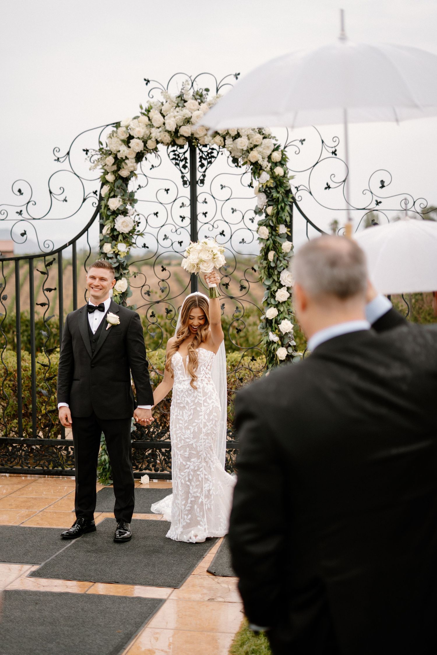 Bride and groom hold hands at outdoor floral arch ceremony while officiant speaks on overcast day.
