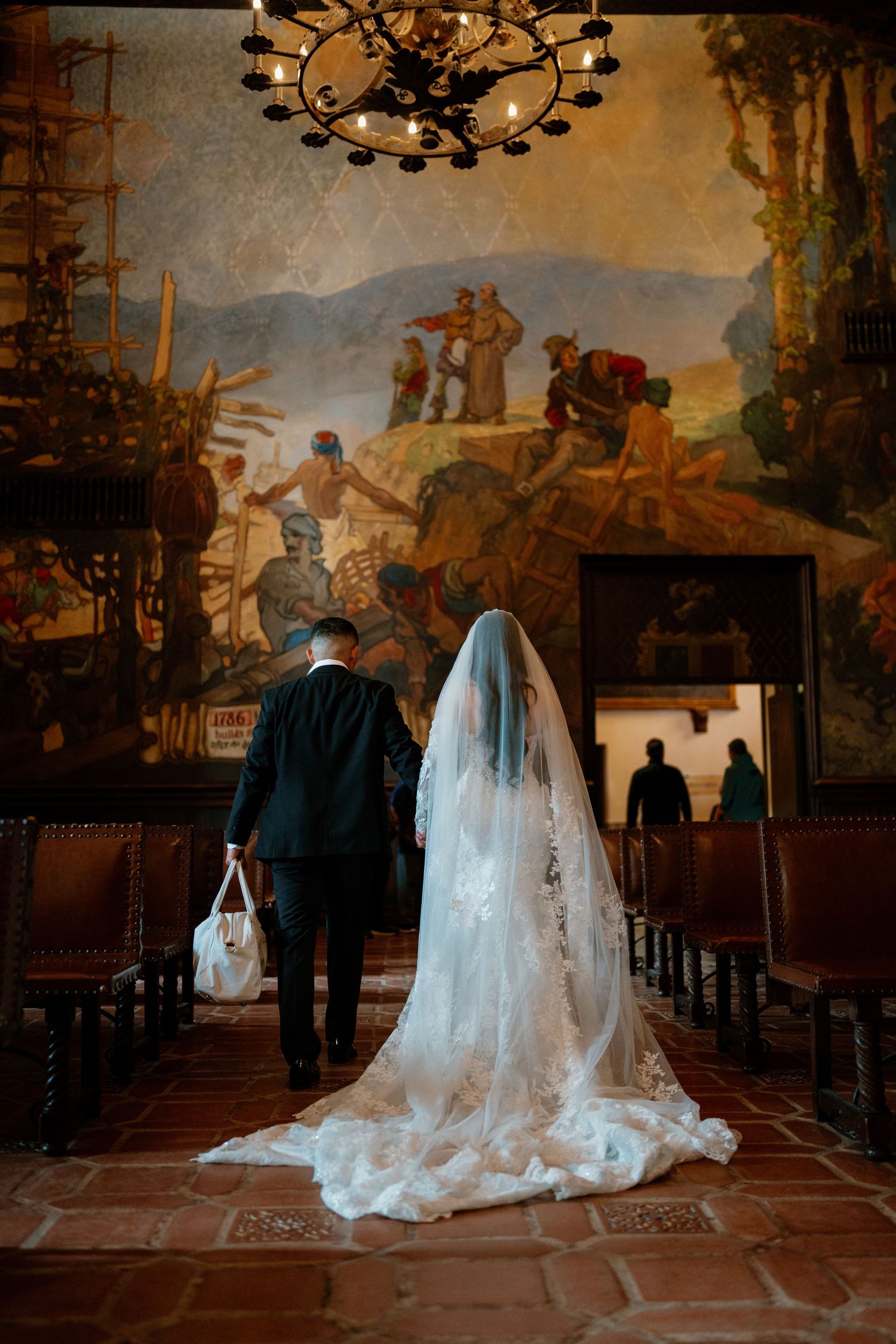 Bride in lace gown with long veil walks with escort toward mural in historic wedding venue.
