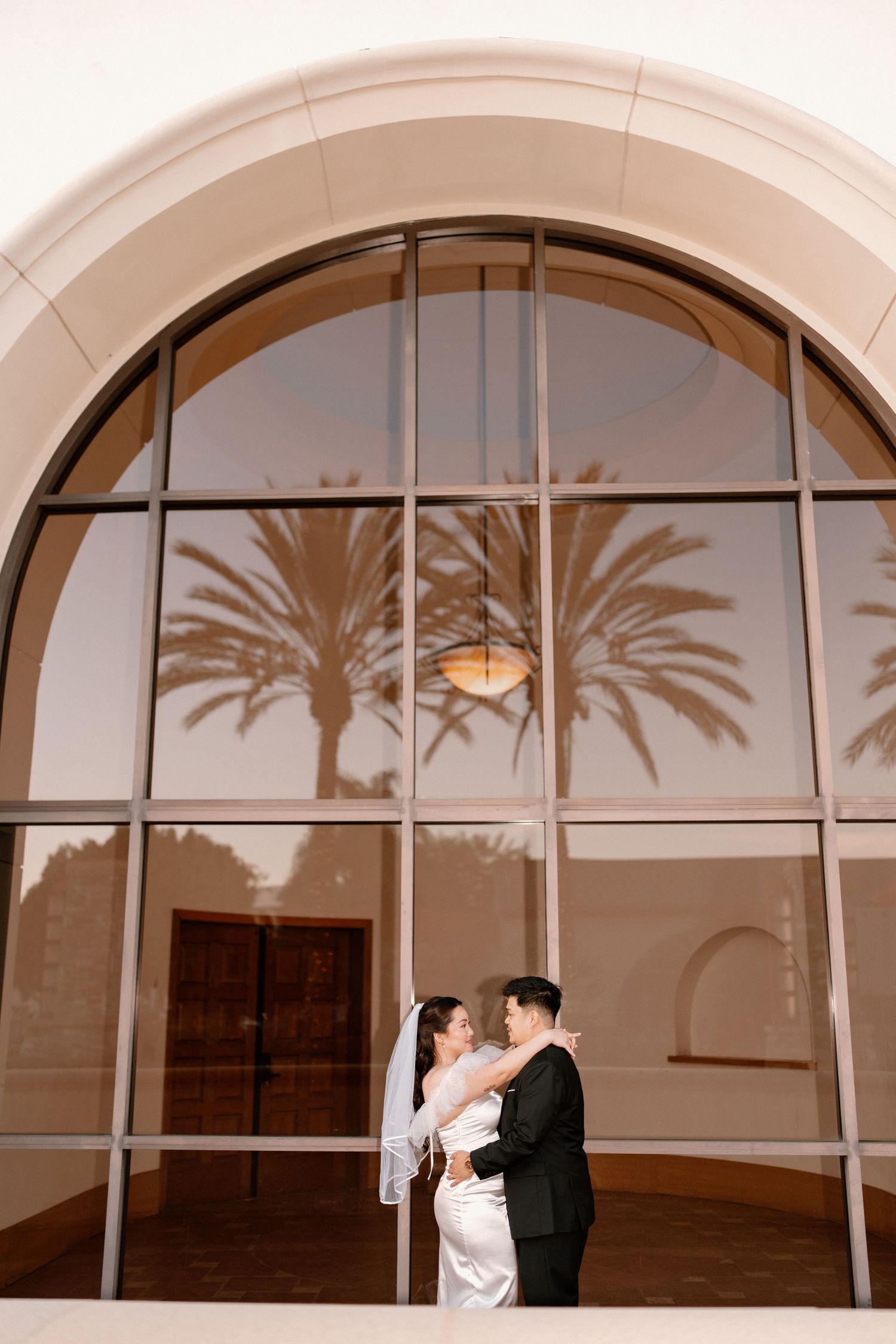 Bride and groom embrace in front of a large arched window reflecting palm trees.